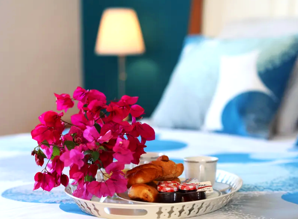Tray with pink bougainvillea flowers, bread, jam, and a small cup on a bed with a blue and white bedding in a bedroom.