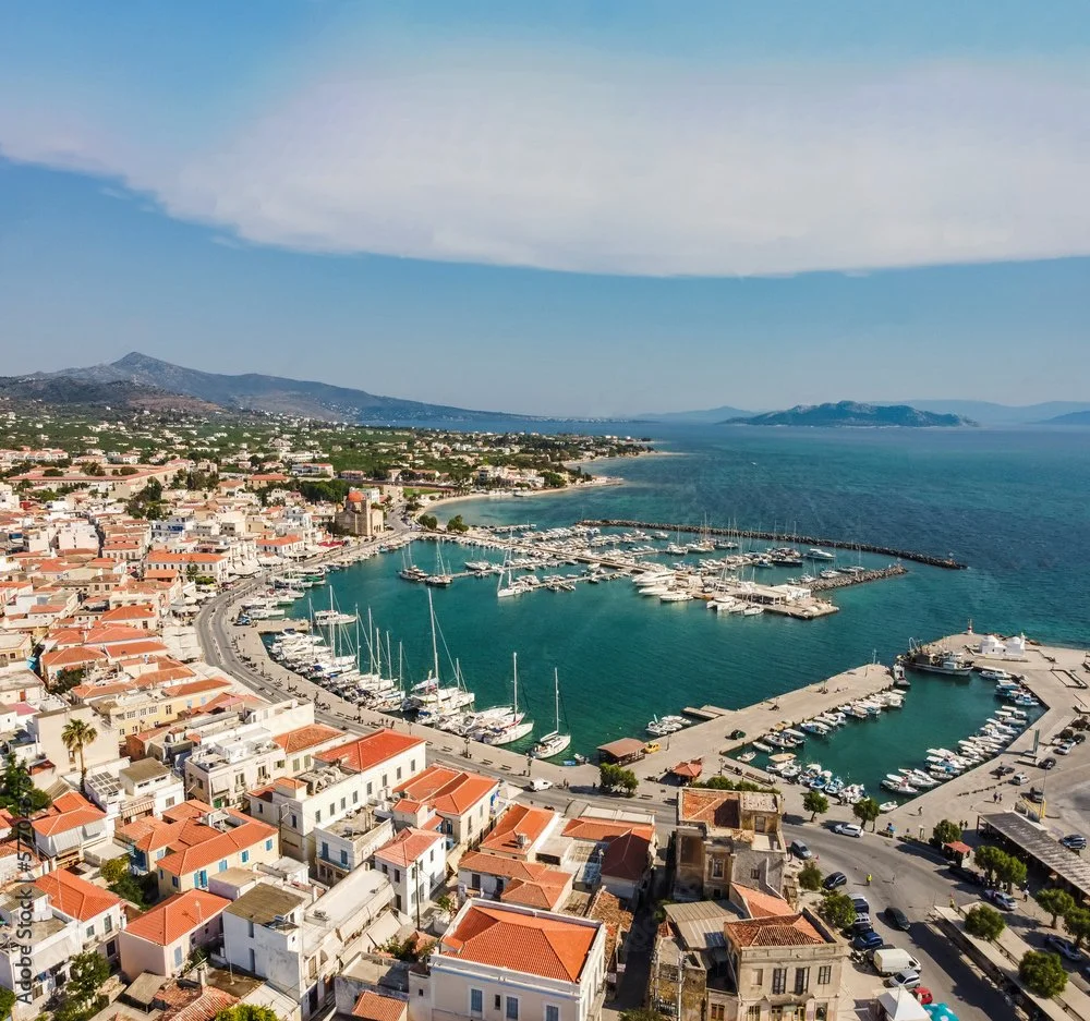 Aerial view of a coastal town with red-tiled roofs, a marina with boats, and a large body of water with islands in the distance.