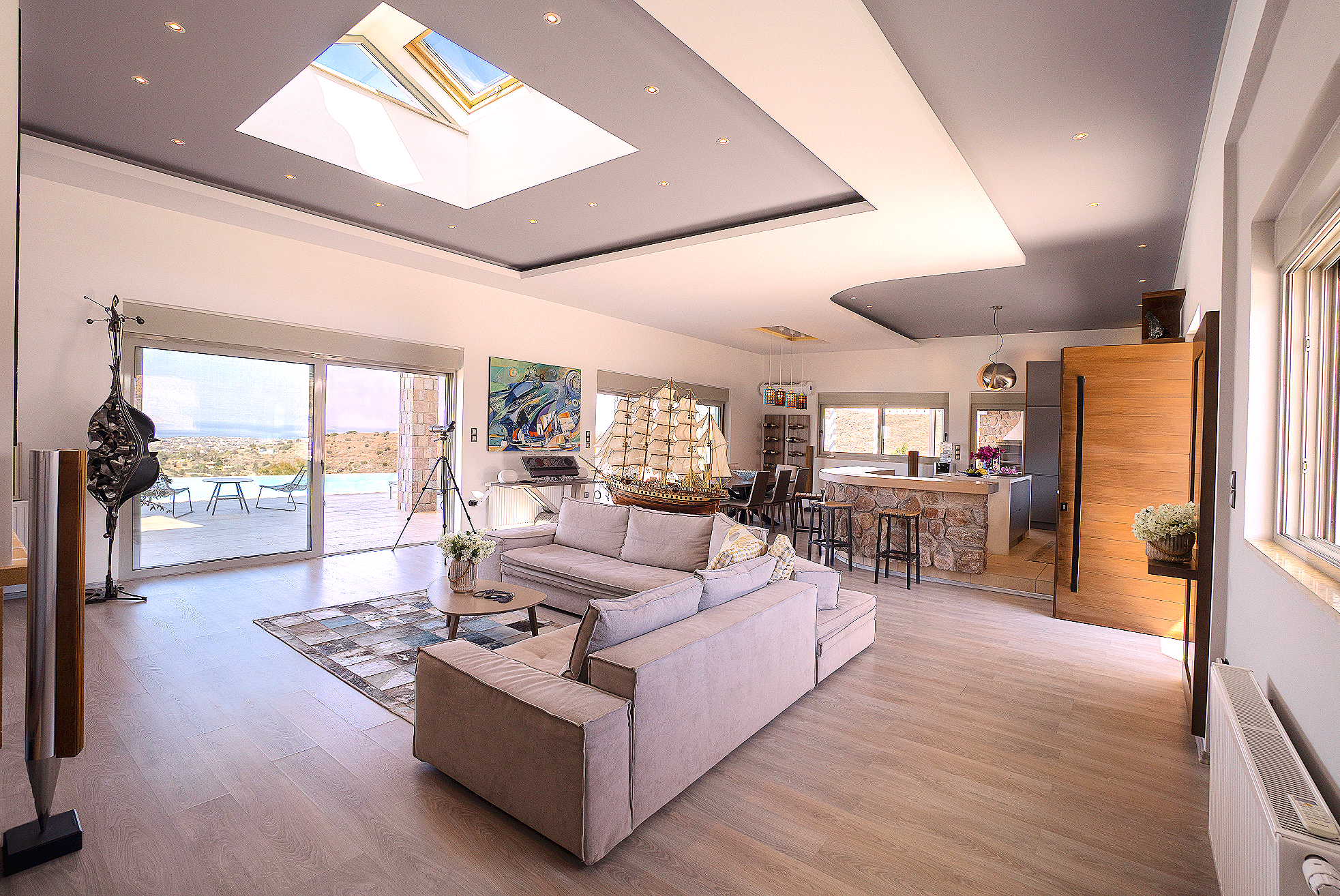 Open-plan living room with beige sectional sofa, nautical model ship, and telescope near sliding glass doors leading to balcony with outdoor seating, view of hills, skylight, and modern kitchen with stone island counter.