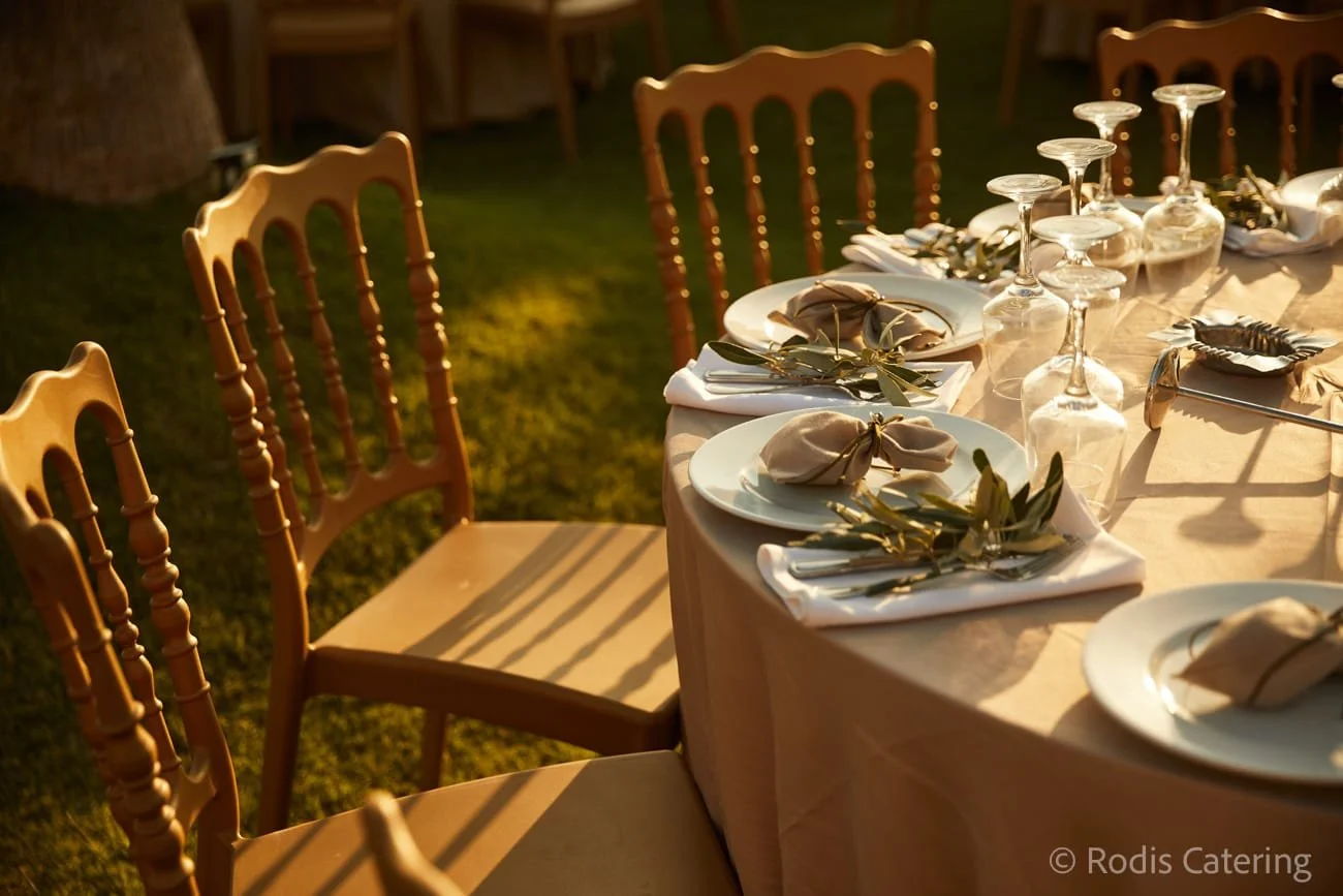 A long outdoor dining table set for a dinner, with white plates, napkins tied with gold ribbons, wine glasses, and silverware, illuminated by warm sunlight and surrounded by wooden chairs.