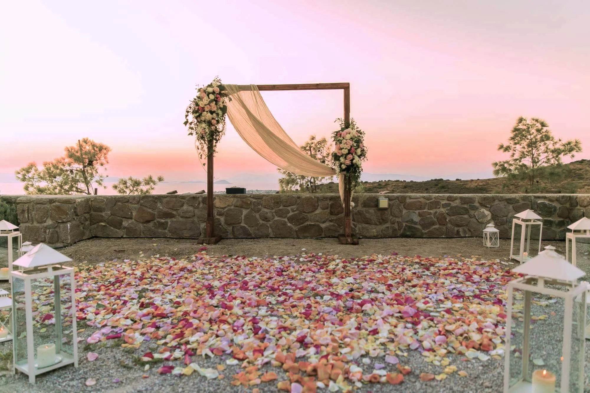 Outdoor wedding ceremony setup with an arch decorated with flowers and fabric, surrounded by lanterns and rose petals on the ground at sunset.