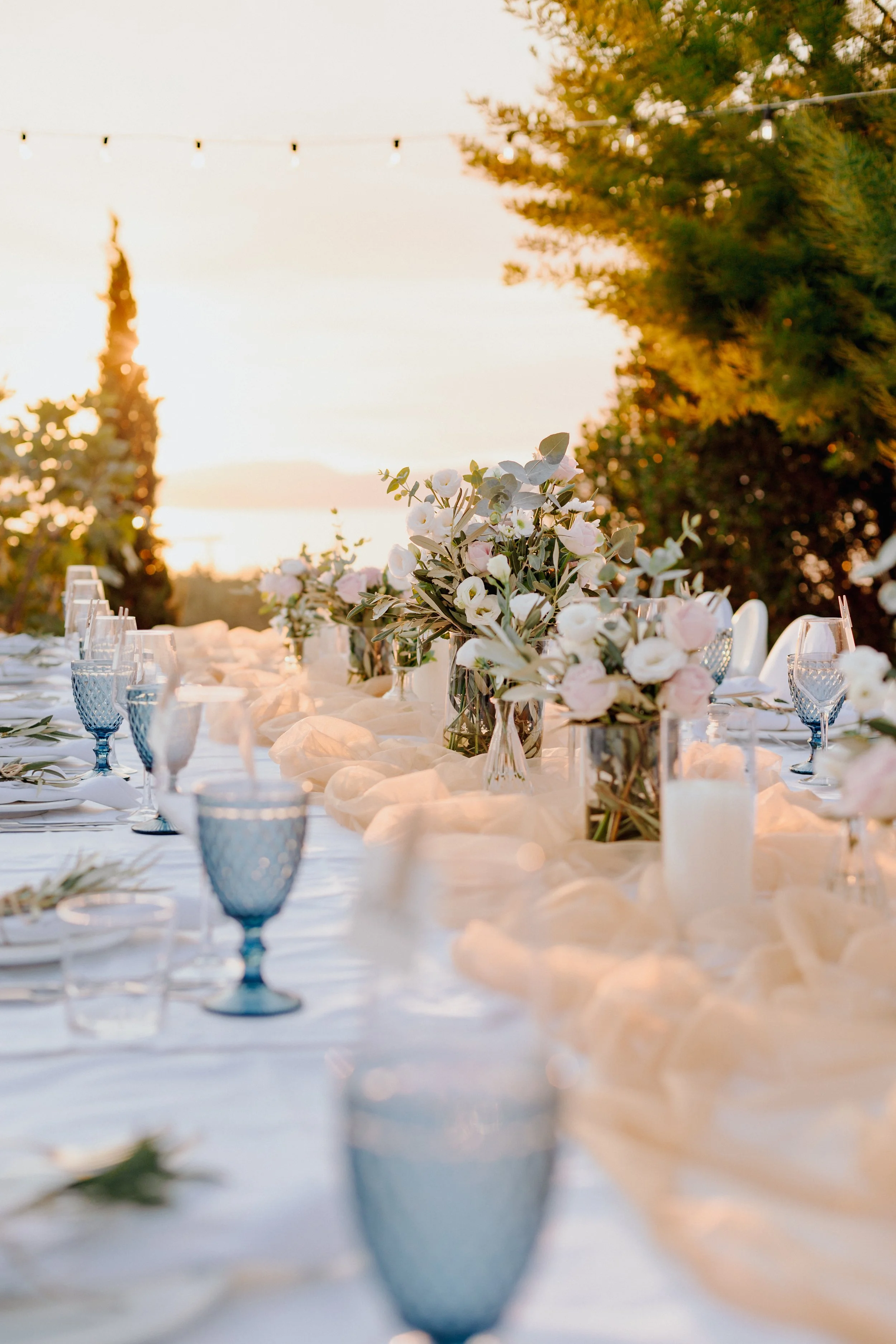 Elegant outdoor table setting at sunset with white tablecloth, floral centerpieces, blue glassware, and candle lanterns, surrounded by trees.
