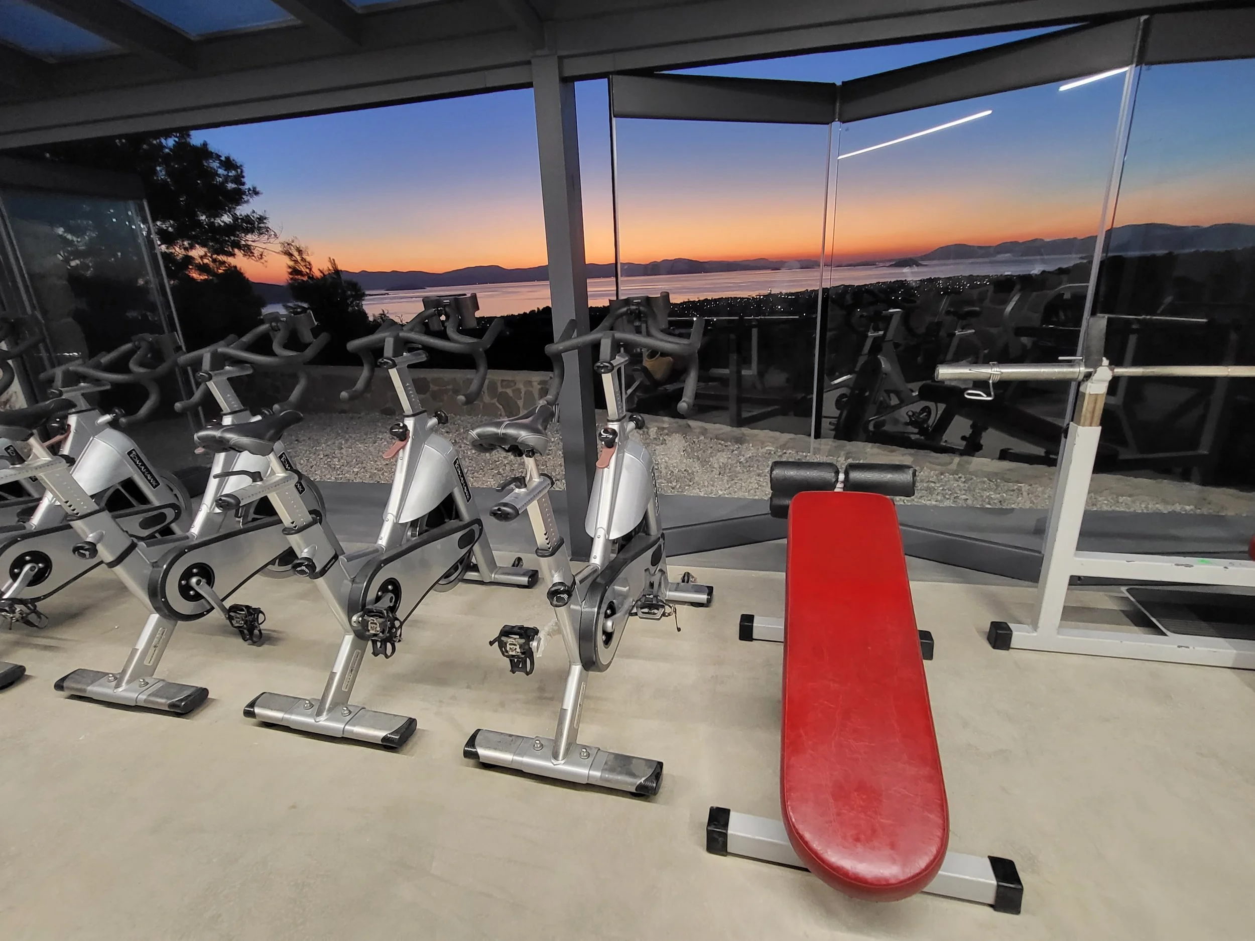 Indoor gym with exercise bikes and weight bench, overlooking a sunset view of mountains and water through large windows.