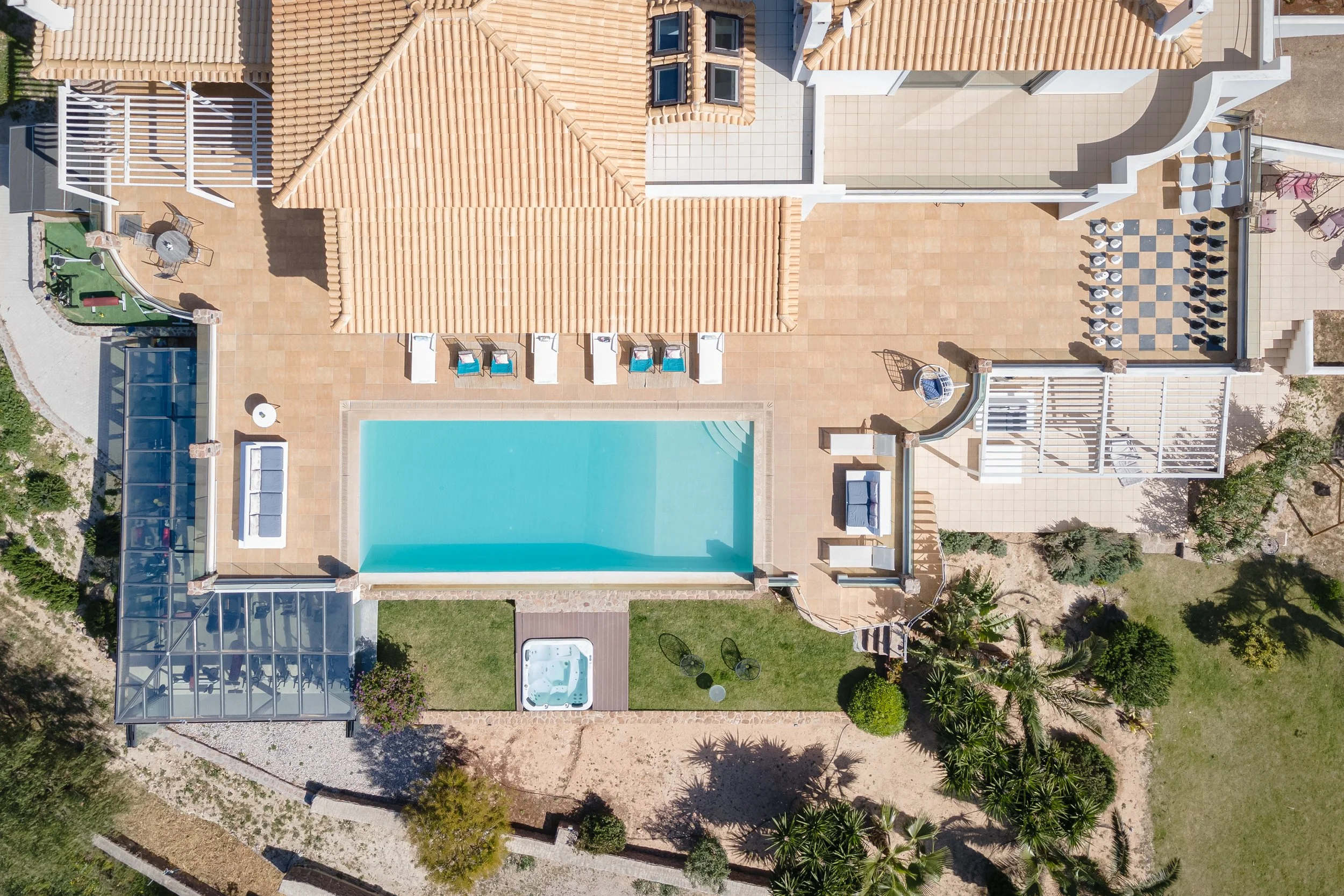Aerial view of a luxurious backyard with a rectangular swimming pool, hot tub, outdoor seating area, and a covered patio with tiled flooring. The yard is landscaped with plants and trees, and the house features a terracotta tiled roof.