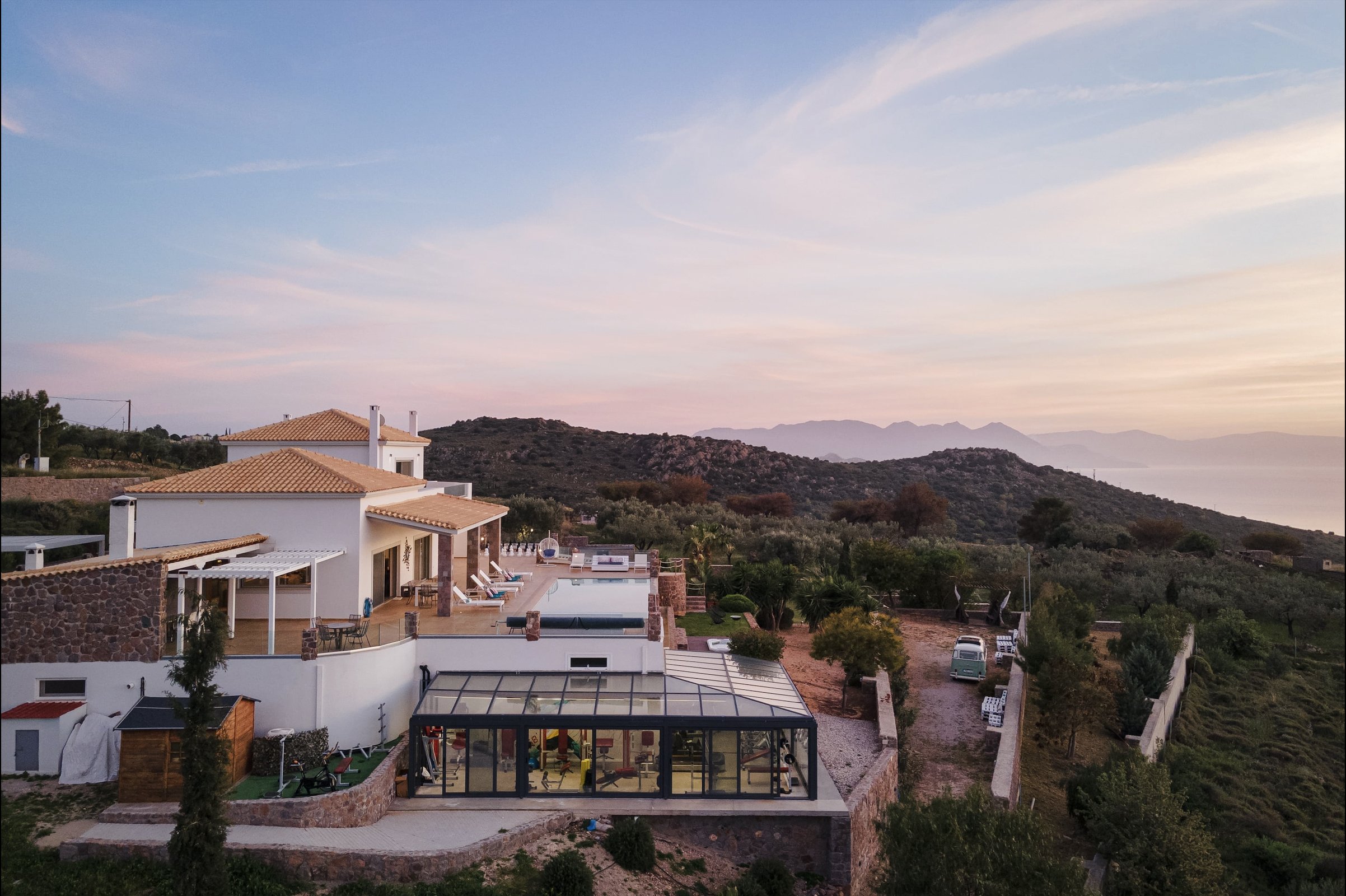 Luxury villa with white walls, red-tiled roofs, and outdoor pool, set amid hilly landscape with mountains and ocean in the background during sunset.