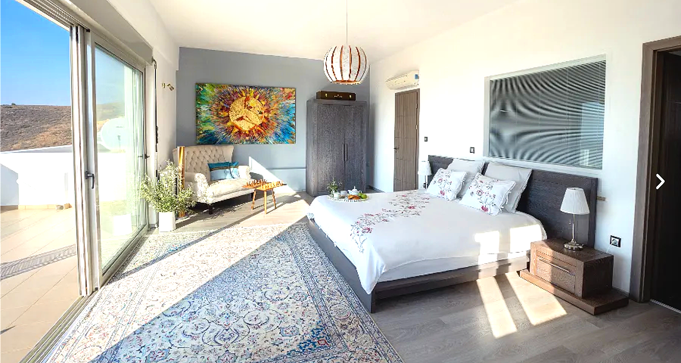 A bright hotel bedroom with a large bed, nightstand with lamp, a sitting area with a small sofa, artwork on the wall, and glass sliding doors leading to a balcony with a view of the outdoors.