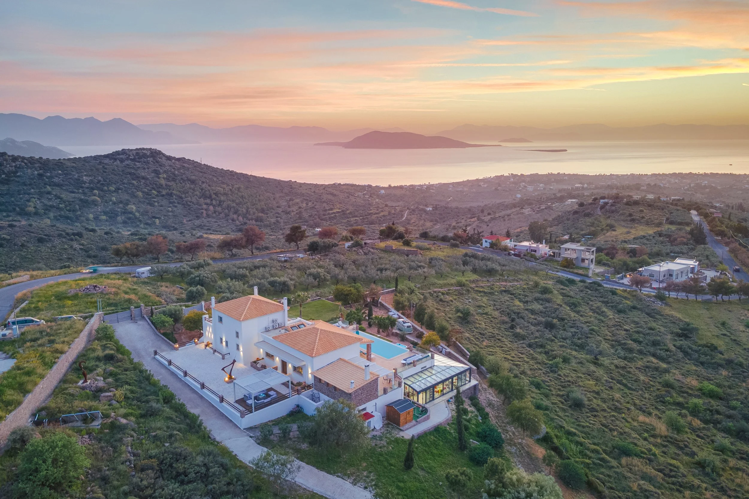 Aerial view of a large white house with terracotta roof tiles, a swimming pool, and outdoor seating, situated on a hillside overlooking a coast at sunset with mountains and water in the distance.