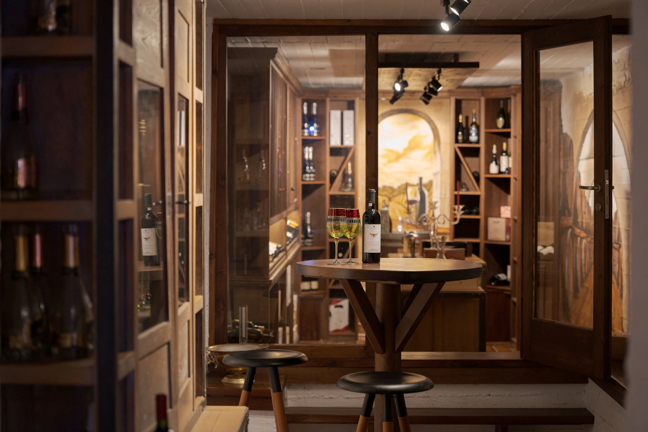 A cozy wine room with glass doors, wooden shelves stocked with wine bottles, a small round table with a bottle of red wine and two wine glasses, and two bar stools. Warm lighting highlights the wood interior and a landscape painting on the wall.