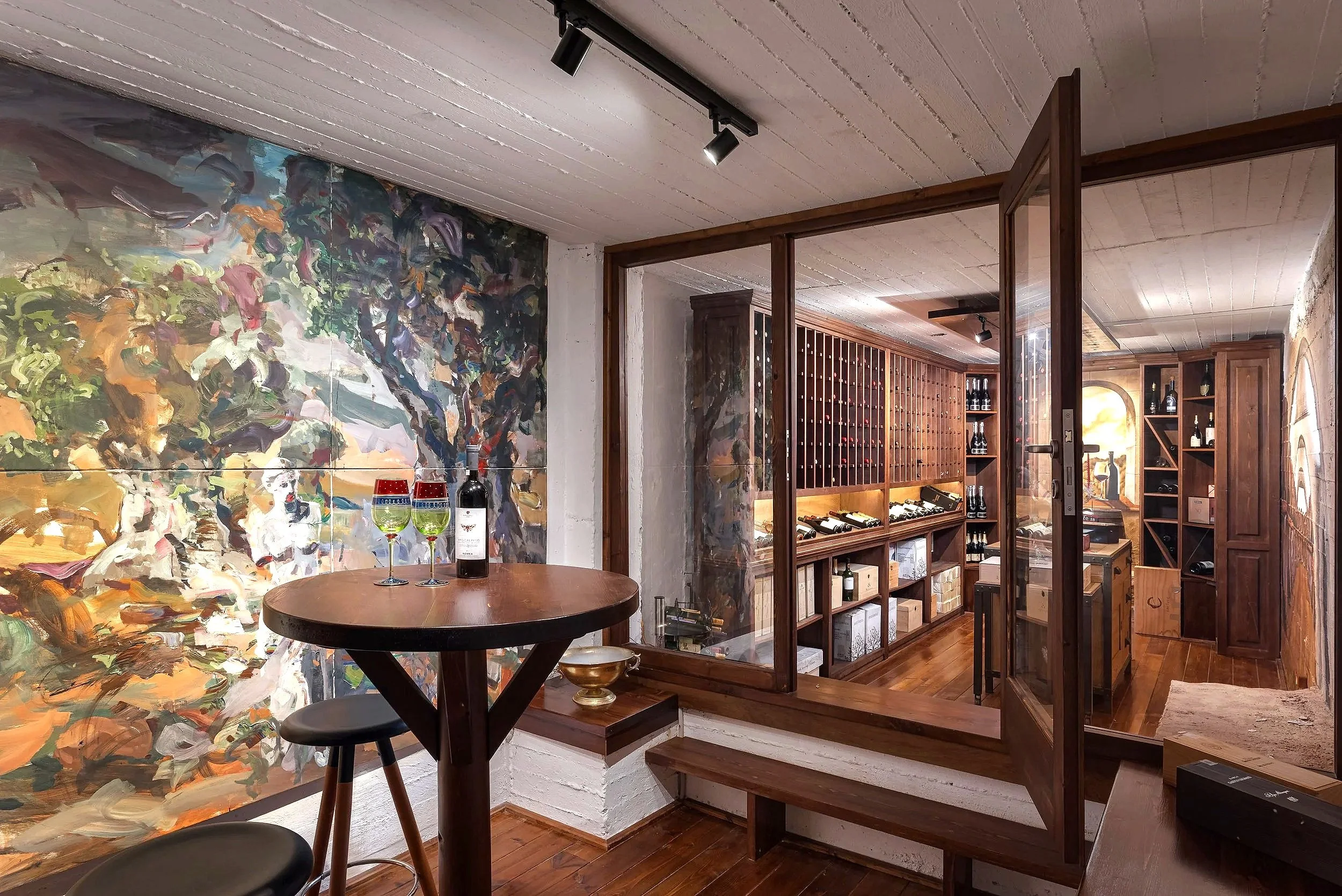 A cozy wine cellar with wooden shelves filled with wine bottles, a small wooden table with two glasses of white wine, and a colorful abstract painting on the wall. The cellar is separated by glass doors and has warm lighting.