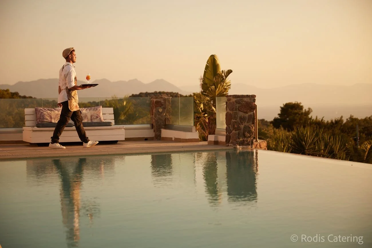 A waiter walks along a poolside patio at sunset, carrying a drink and a tray, with mountains in the distance and a lounge area with pillows and plants nearby.