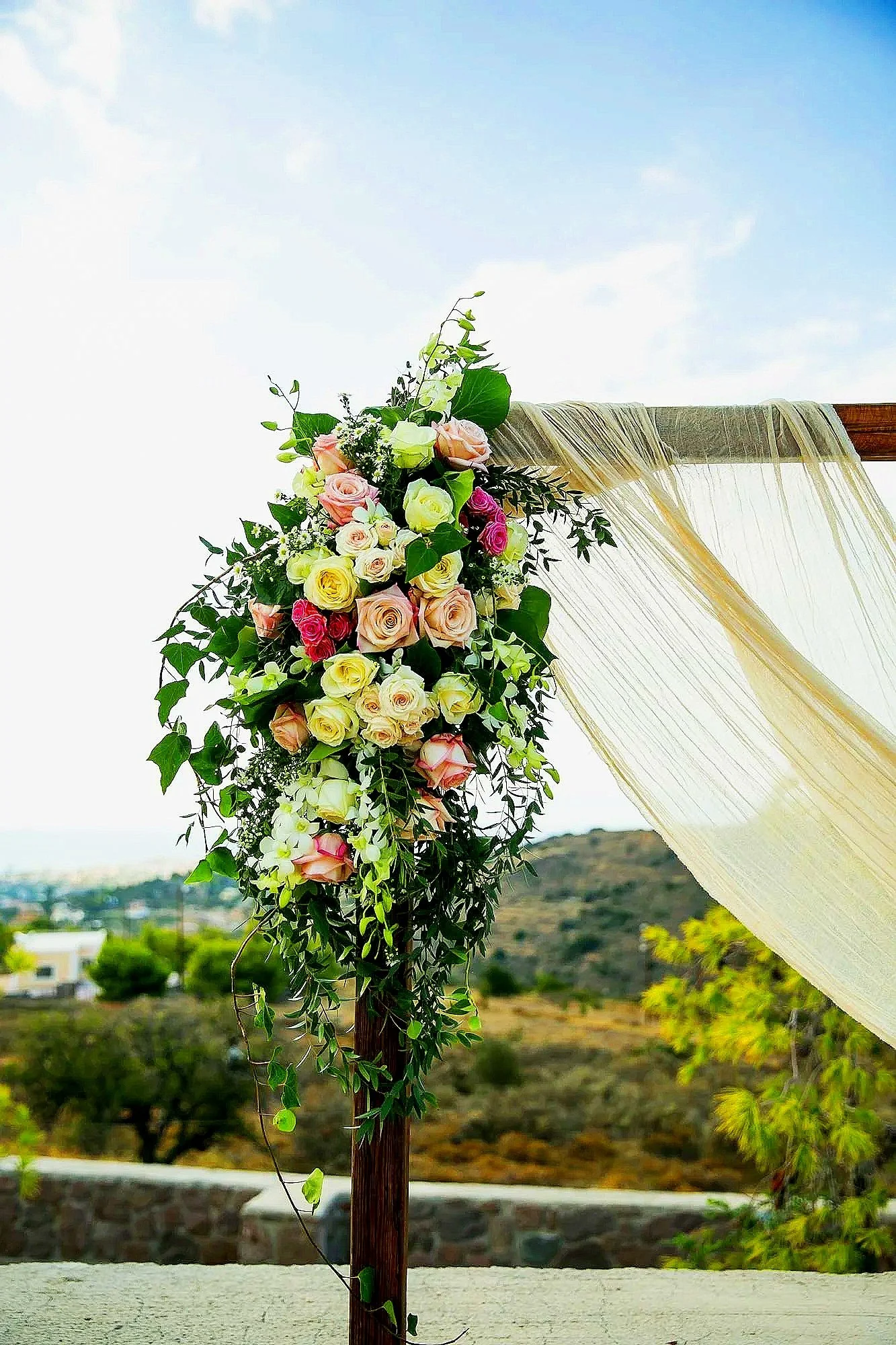 A floral arrangement with pink, yellow, and white roses and greenery attached to a wooden post with sheer fabric draped on it, set outdoors against a scenic landscape.