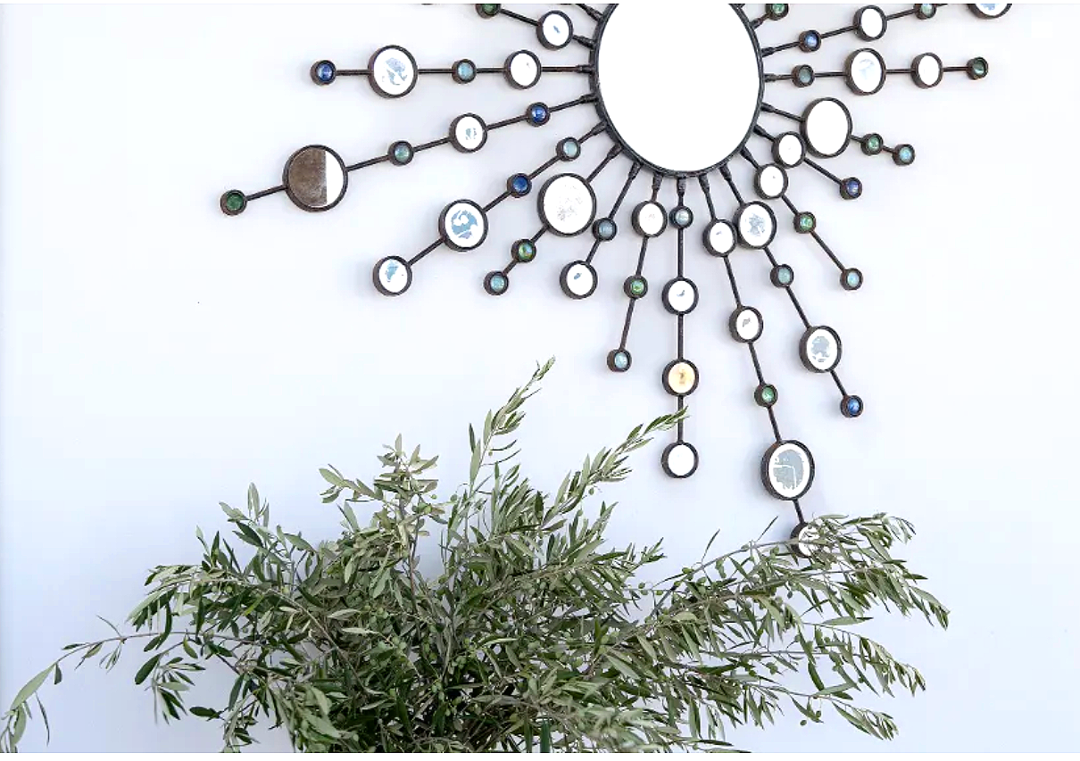 Decorative wall mirror with a sunburst design, with branches extending outward, ornaments, and mirrors, placed above a green leafy plant against a light-colored wall.