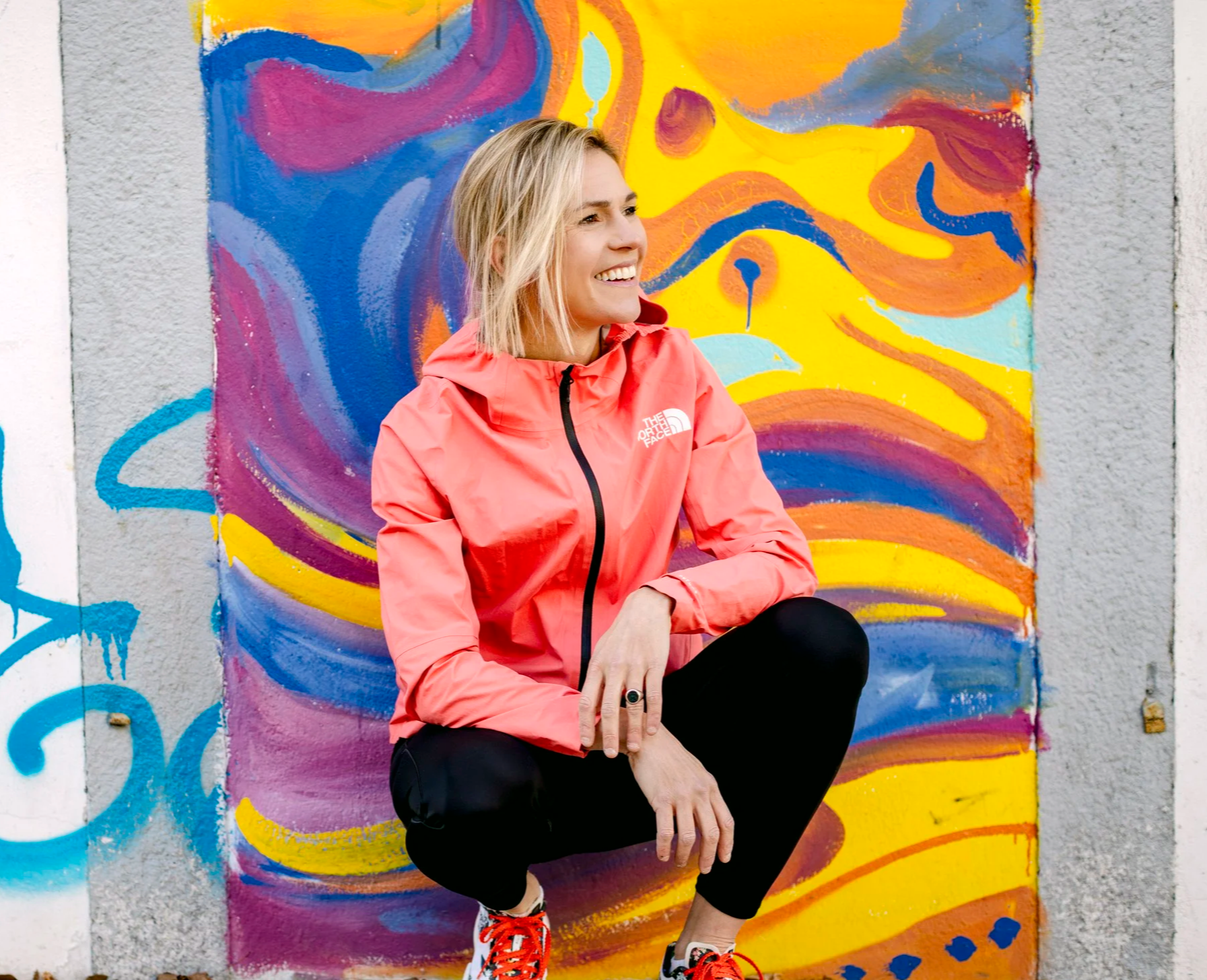 A woman with blonde hair, wearing a coral pink North Face jacket and black pants, is crouching and smiling in front of a colorful abstract mural on a wall.