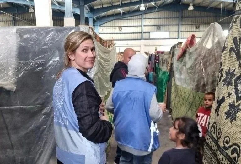 A woman standing inside a shelter with children and other adults, some wearing jackets and hoodies, around covered structures in an indoor space with a metal roof and beams.