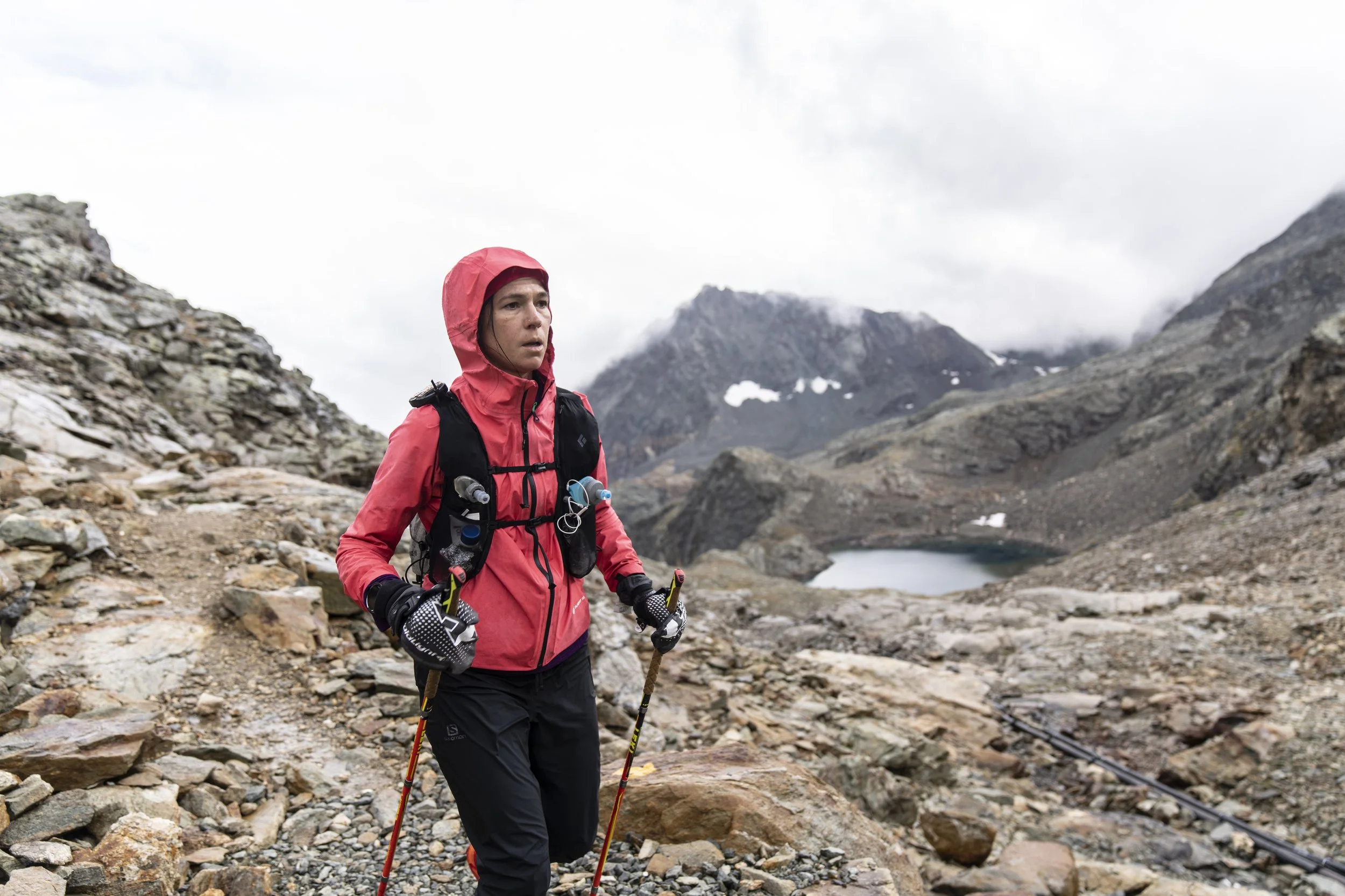 Stephanie Case in a red jacket and black pants hiking with trekking poles in a rocky, mountainous landscape with cloudy skies, a lake in the background, and snow patches on the mountains, days into the 450km Tor des Glaciers course.