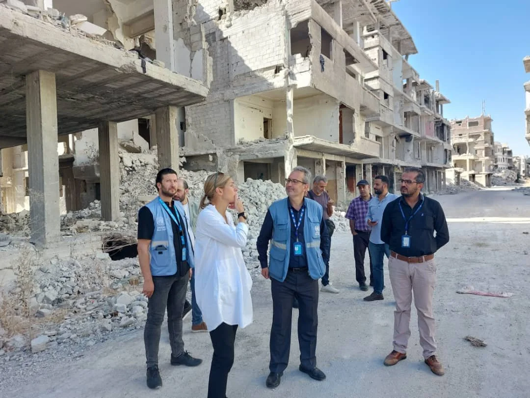 Group of people, including Stephanie Case in white and men wearing blue vests, standing in front of damaged buildings and rubble in Yarmouk camp in Syria.
