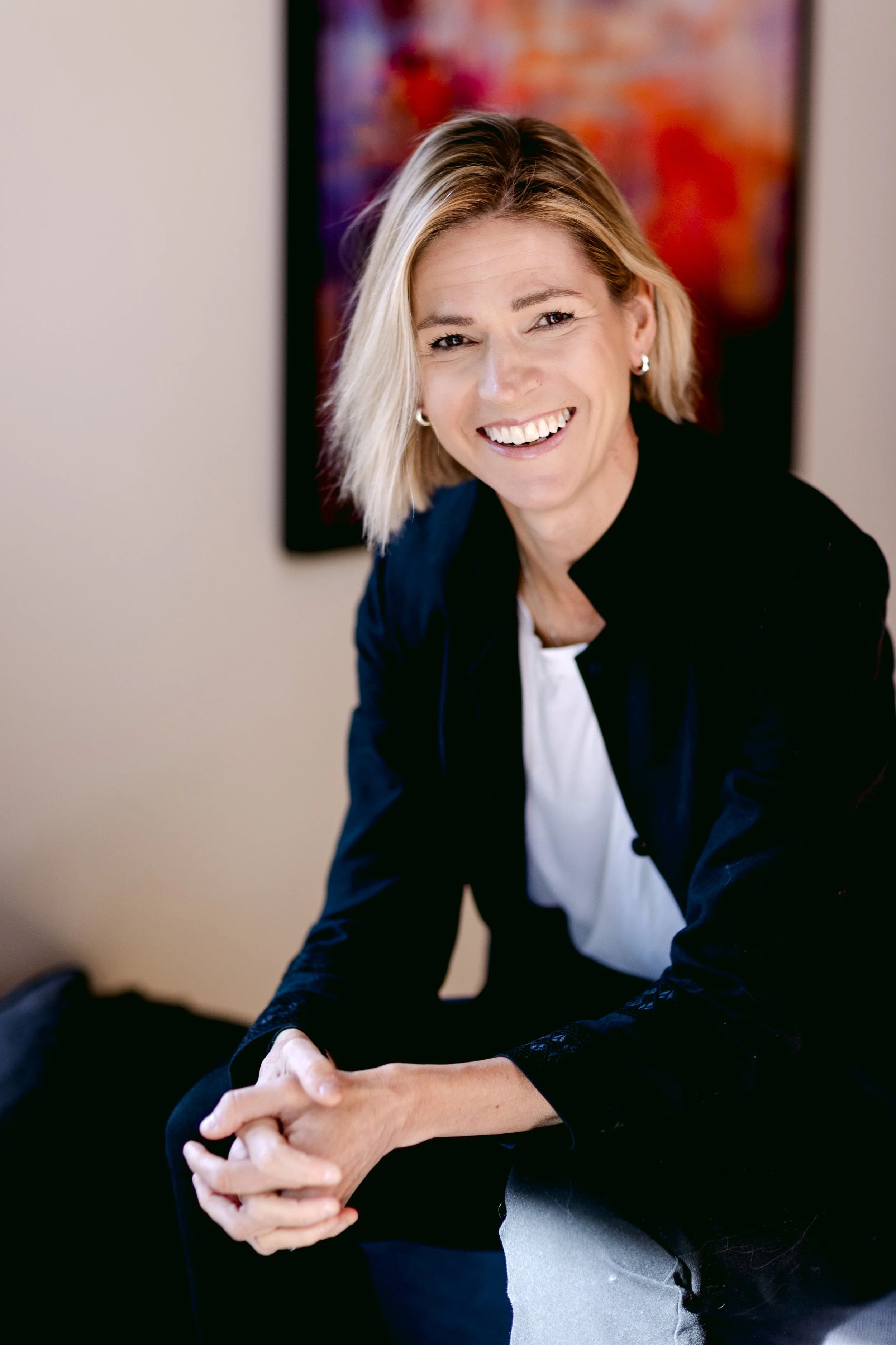 Stephanie Case, smiling and wearing a black blazer over a white top, sitting indoors with a colorful abstract painting in the background.
