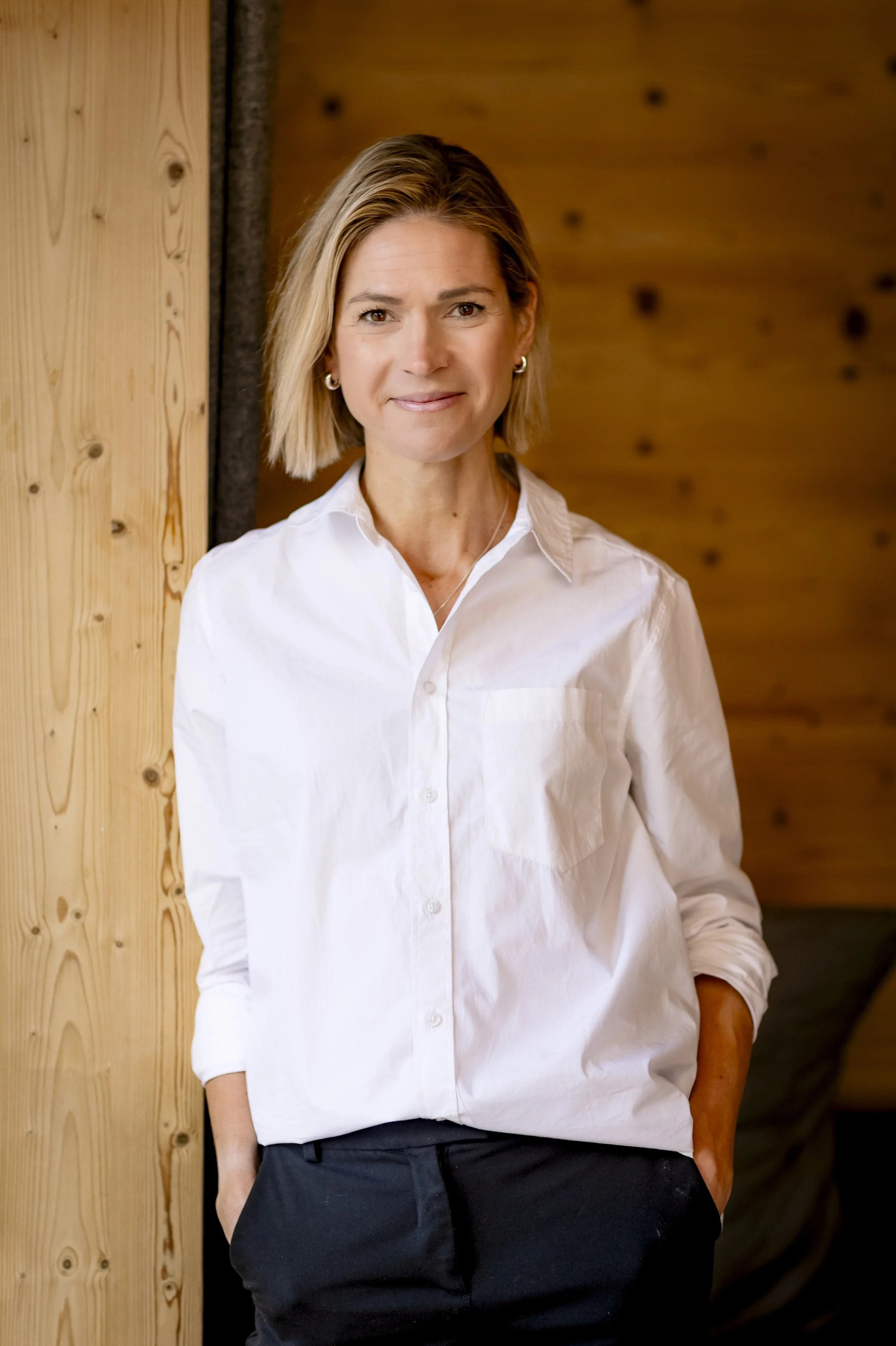 Stephanie Case in a white button-up shirt and black pants, standing indoors with wooden walls and a relaxed expression.
