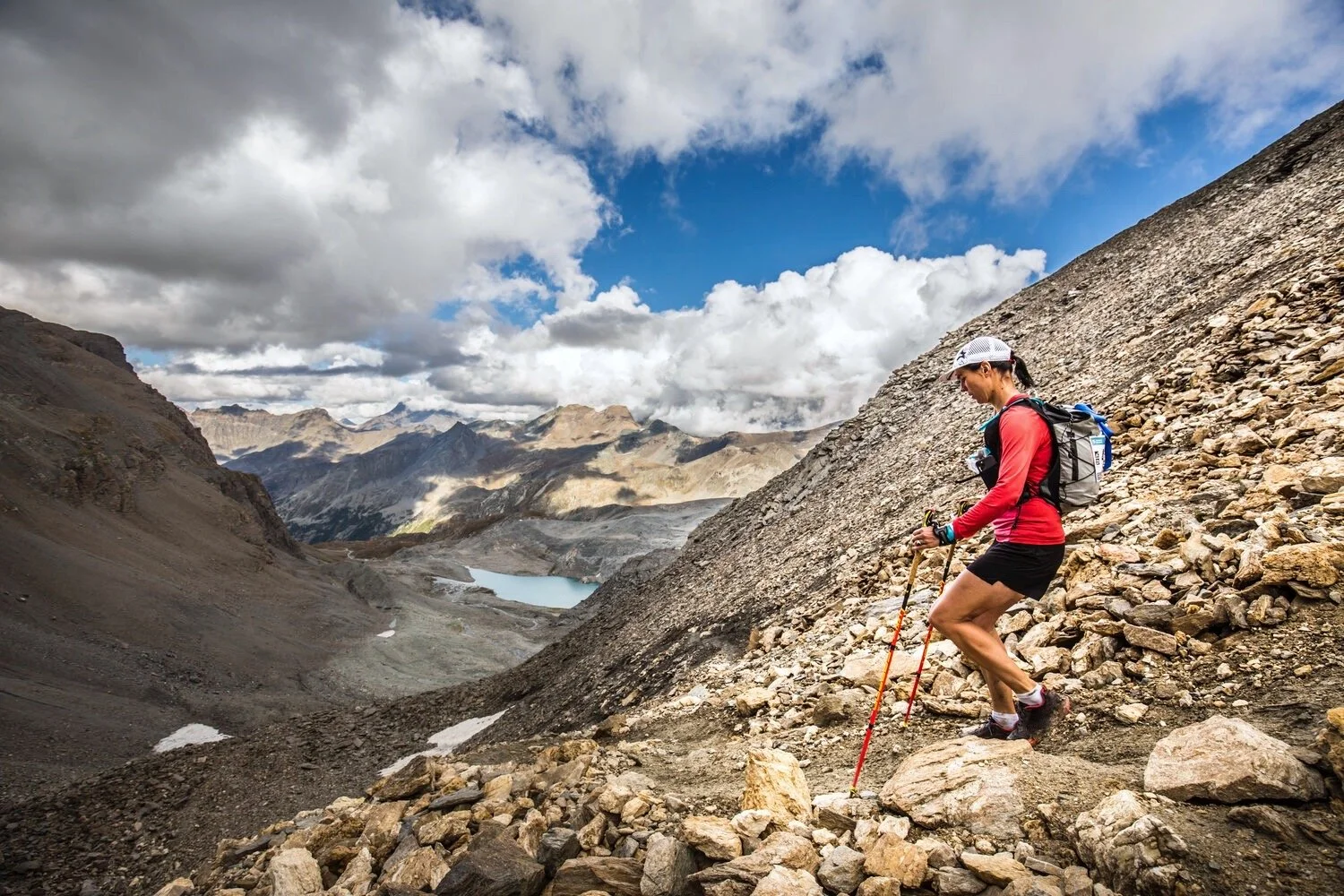 Stephanie Case running down a rocky slope in a mountainous landscape under a partly cloudy sky, carrying a backpack and using trekking poles during the 450km Tor des Glaciers race in 2021.