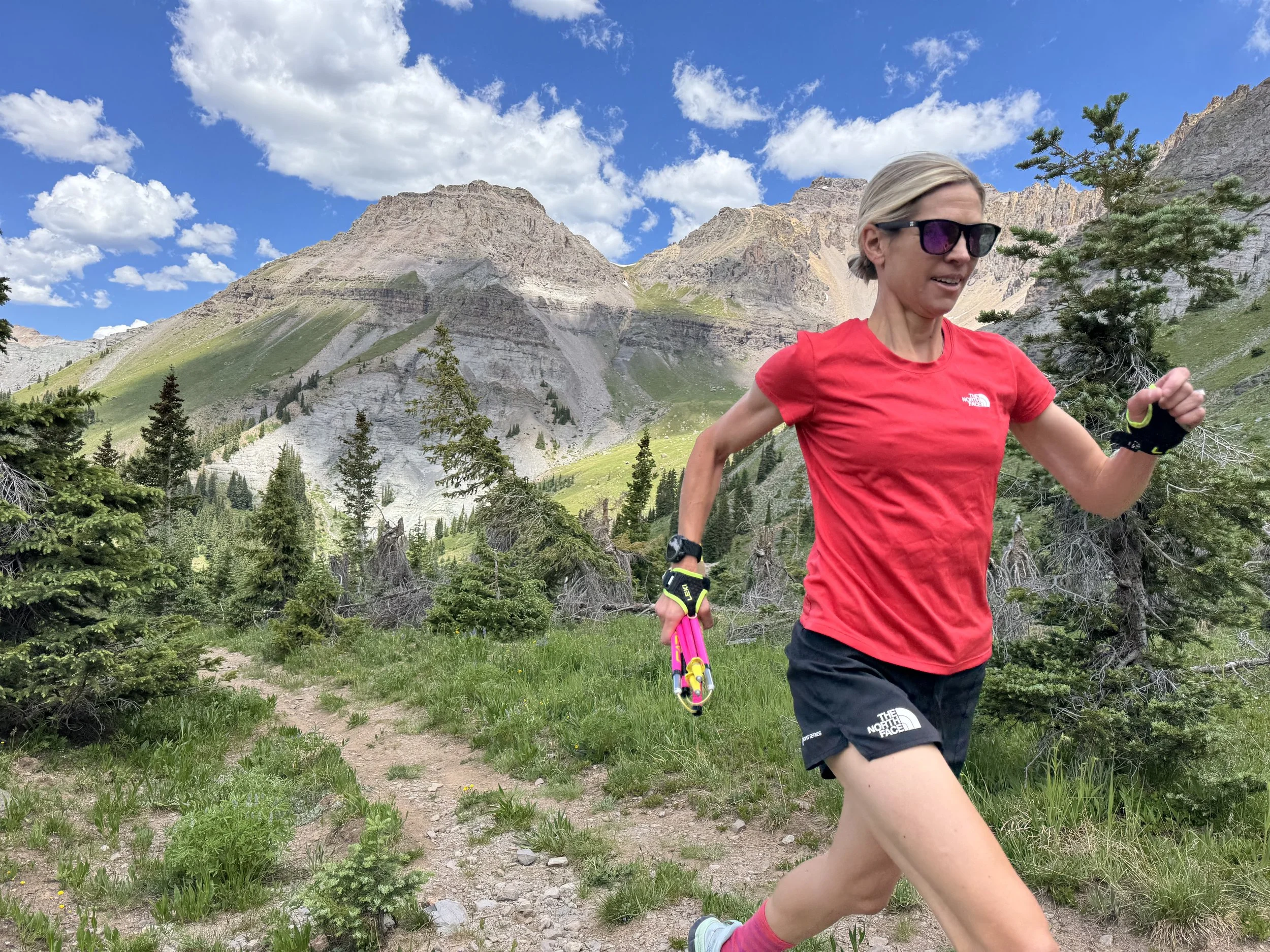 Stephanie Case in red shirt and black shorts running on a mountain trail surrounded by trees in Colorado, with mountains and a blue sky with clouds in the background, just days before the Hardrock Hundred Endurance Run.