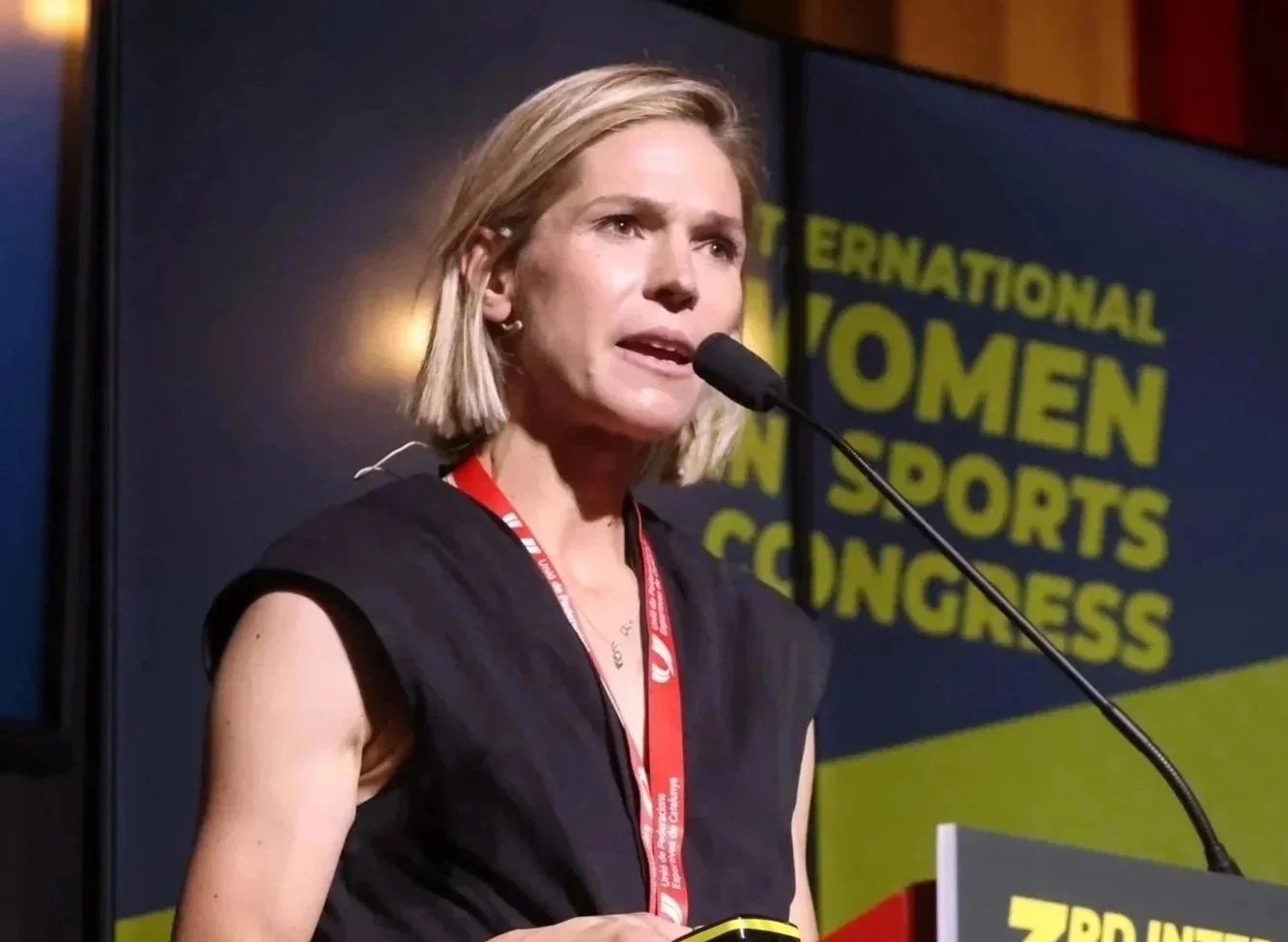 A woman speaking at a conference with a sign behind her that reads "International Women in Sports Congress."