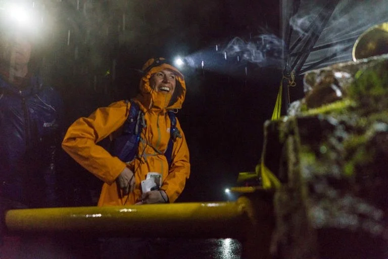 Stephanie Case in an orange raincoat and headlamp smiles with rain and fog around her while at the yellow gate during the Barkley Marathons.