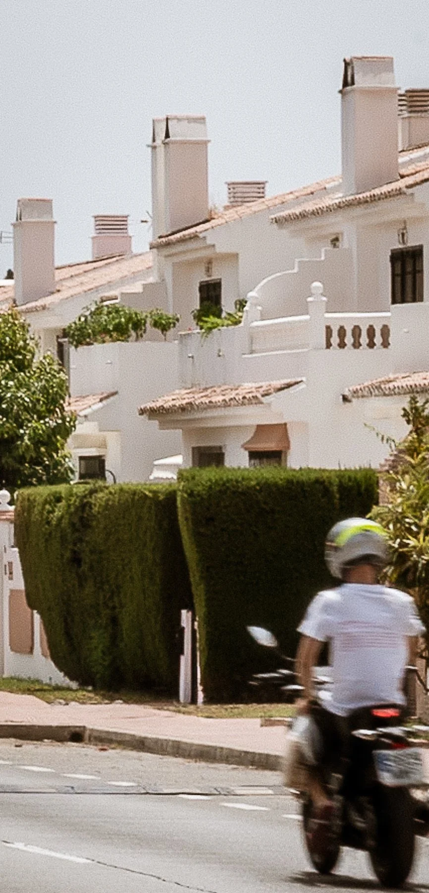 A person riding a motorcycle on a street with a white residential building in the background, which has multiple chimneys, a tiled roof, and a small balcony with decorative railing. There is a large, well-trimmed hedge and some trees visible.