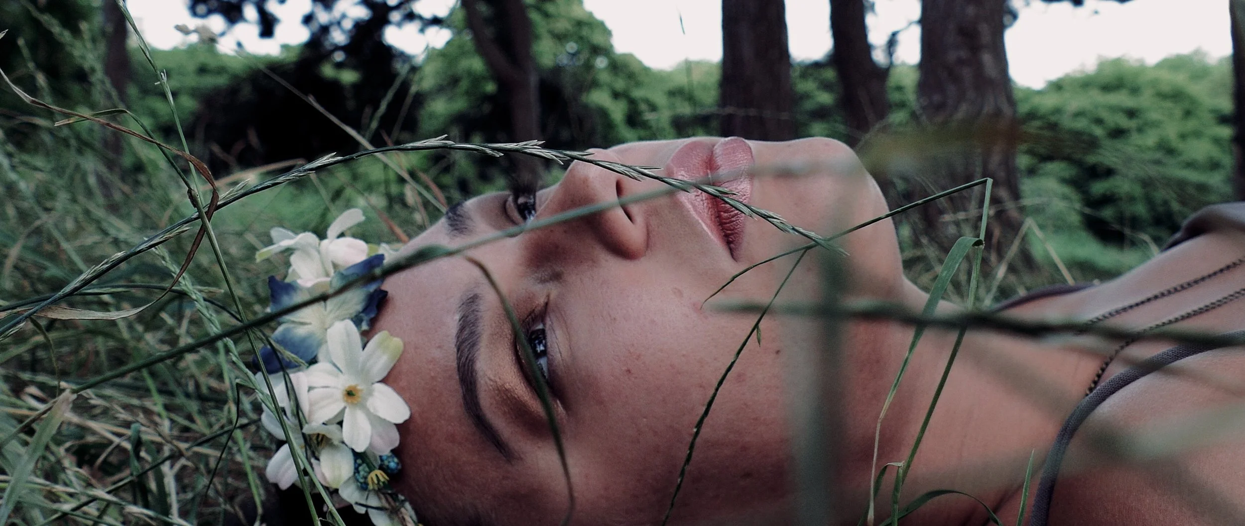 A woman lying down in a grassy outdoor area with trees in the background, wearing a flower crown and a sleeveless top, with her eyes open and looking upward.