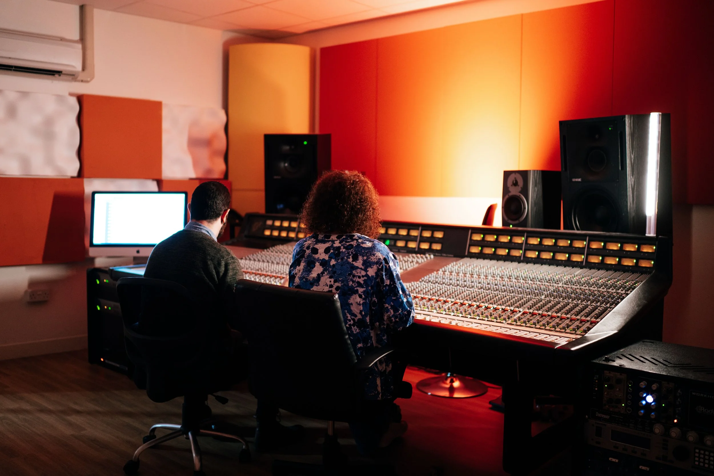 Two people in a recording studio, one has curly hair and the other has short dark hair, working at a large mixing console with a computer monitor, studio speakers, and soundproof walls.