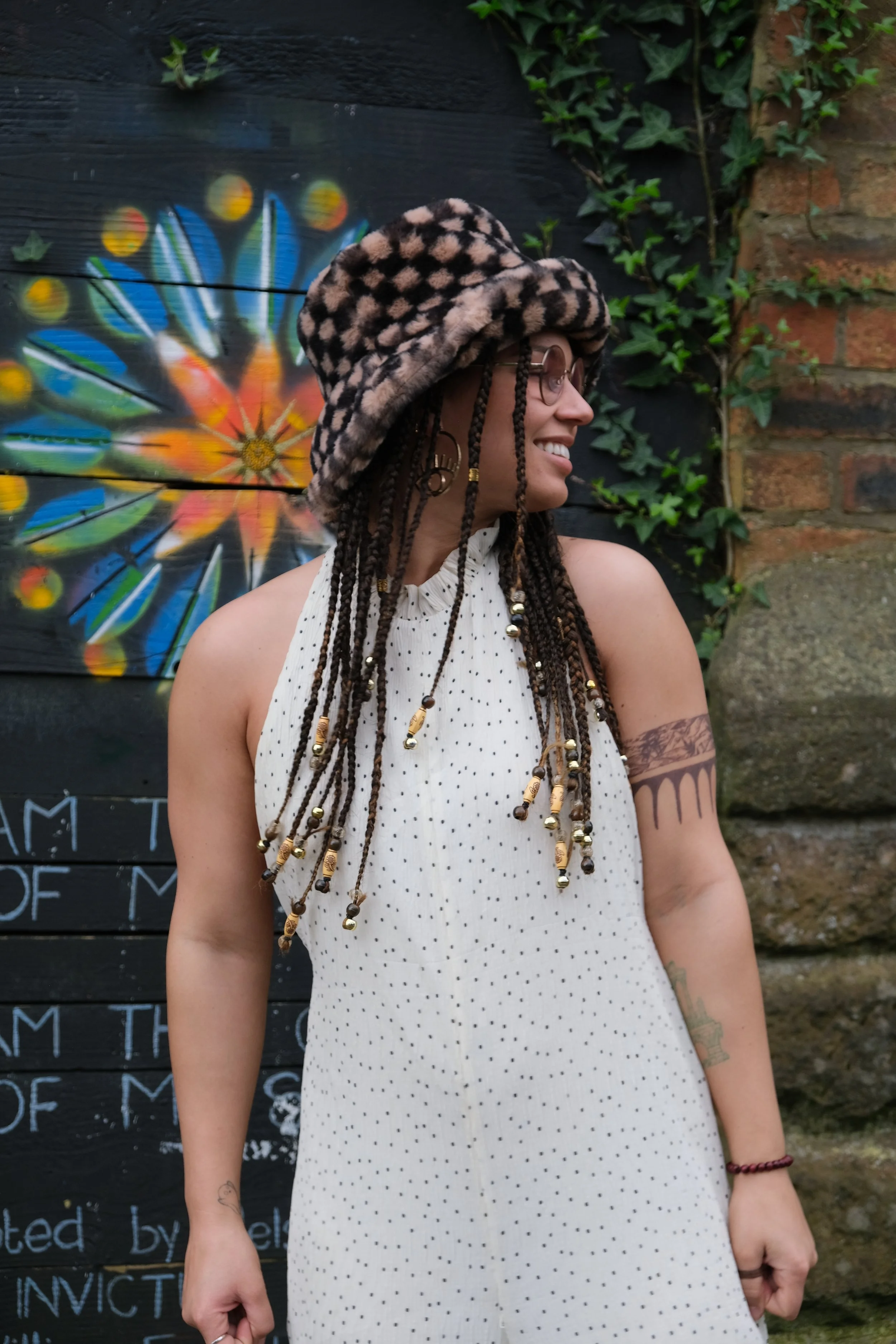 A woman with dreadlocks wearing a leopard print hat and glasses, smiling and looking to the side, standing outdoors against a black wall with colorful graffiti artwork and green ivy.