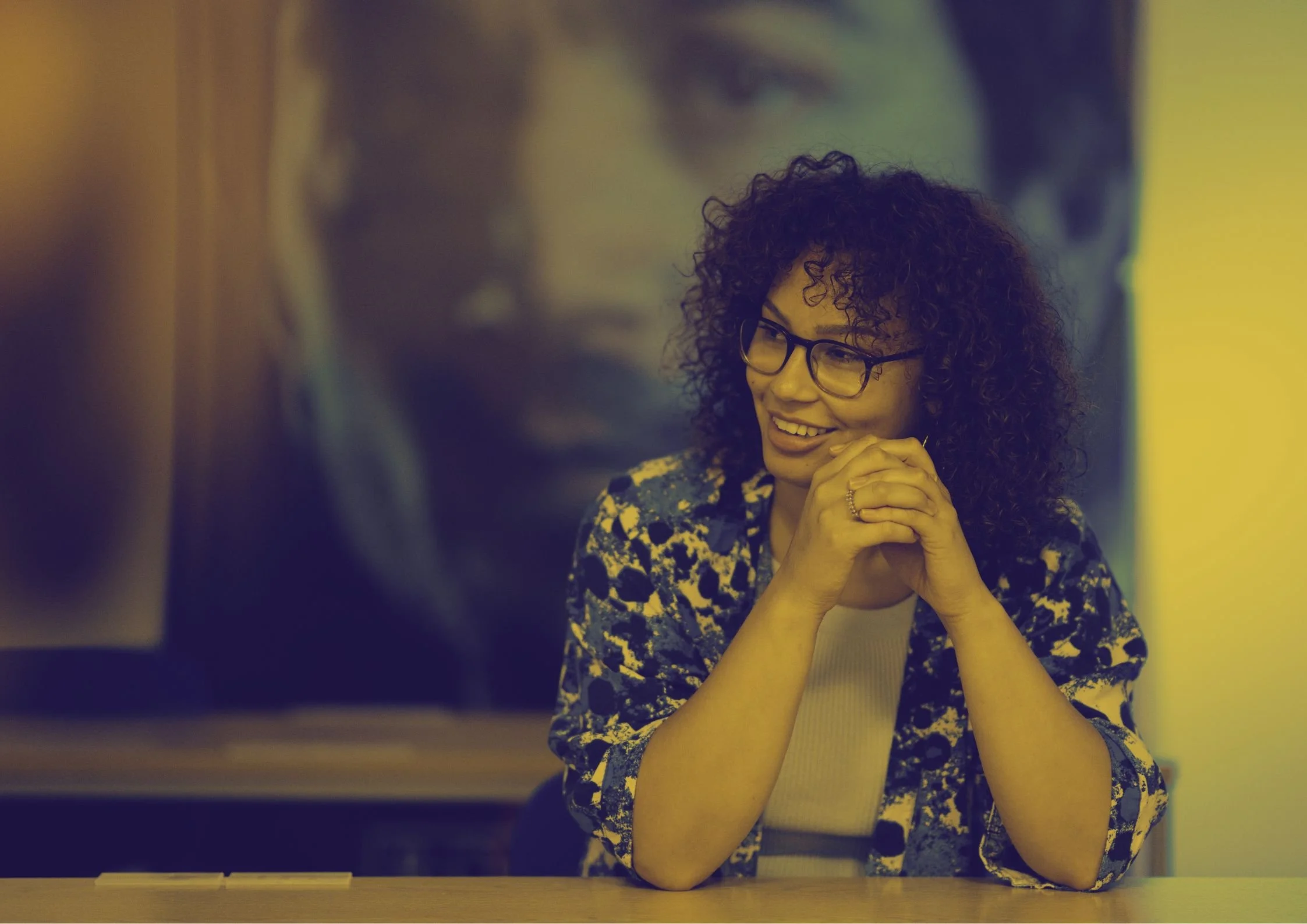 A woman with curly hair and glasses smiling while sitting at a table, clasping her hands near her chin in a well-lit room.