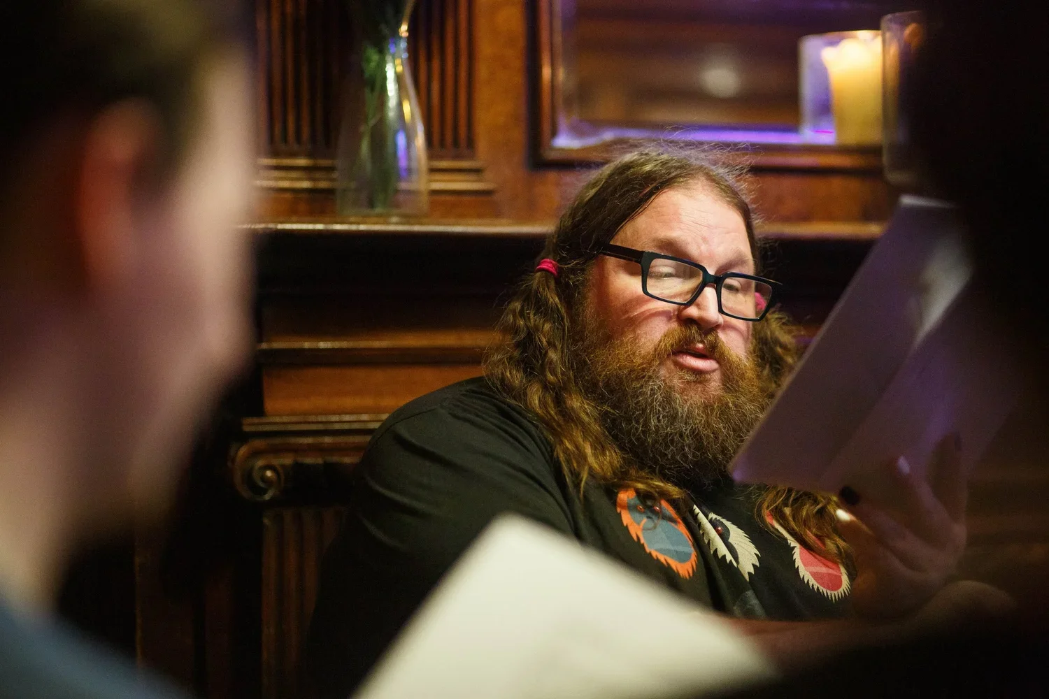 A man with long hair, glasses, and a beard looking at a tablet or phone in a dimly lit indoor setting, with candles and a mirror in the background.
