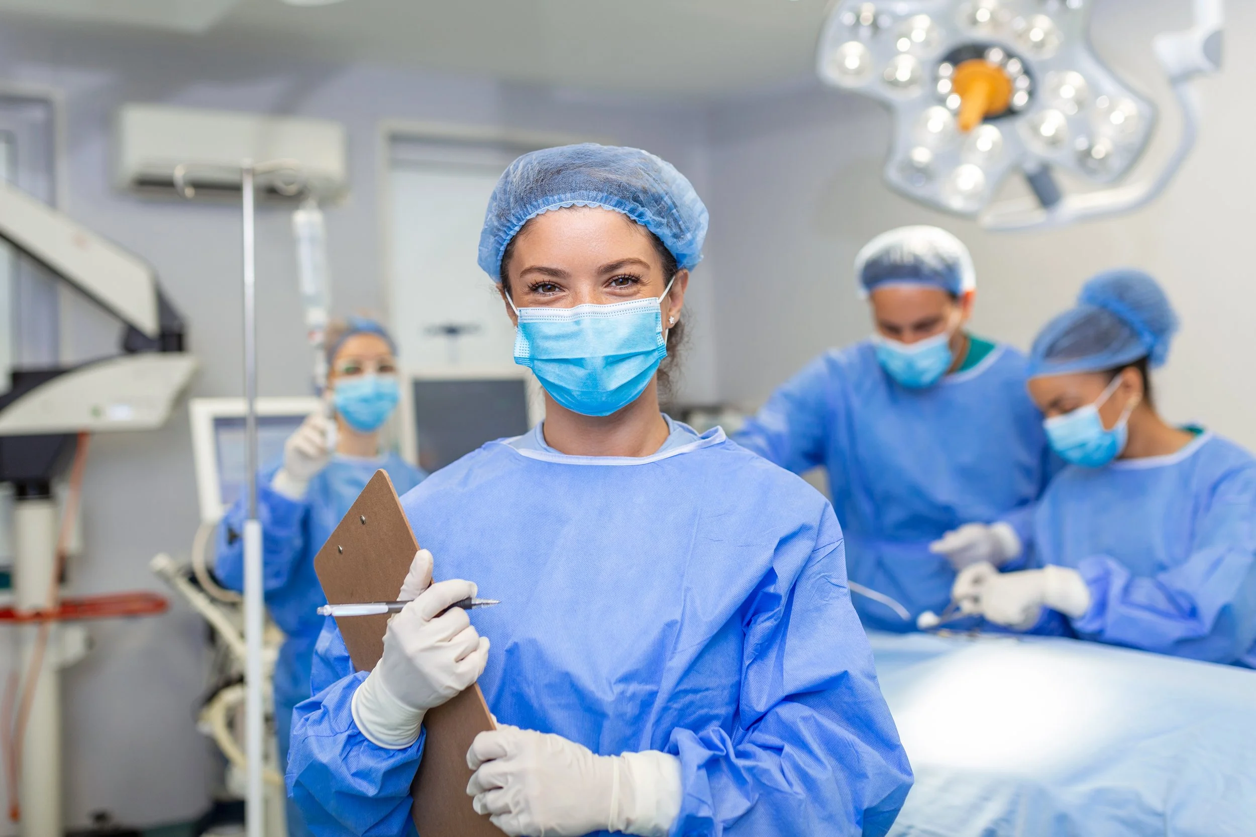 Group of medical professionals dressed in blue surgical scrubs, masks, gloves, and hair covers in an operating room with surgical lights and equipment.
