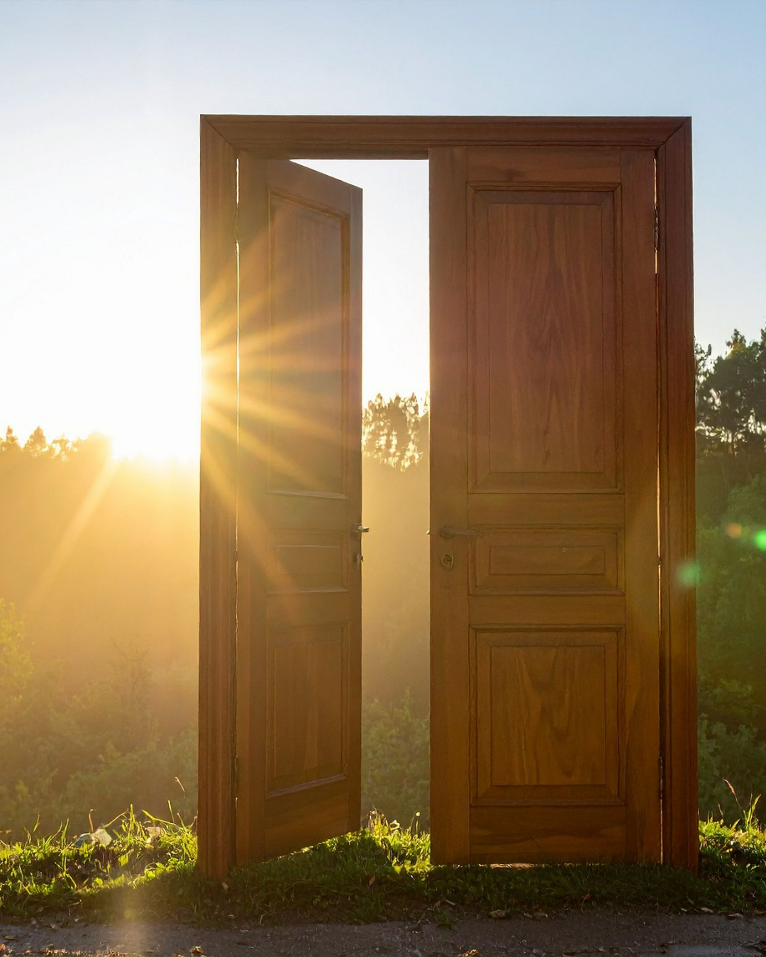 A wooden door standing open on a grassy hill, with sunlight shining through the opening against a backdrop of trees and a clear sky.