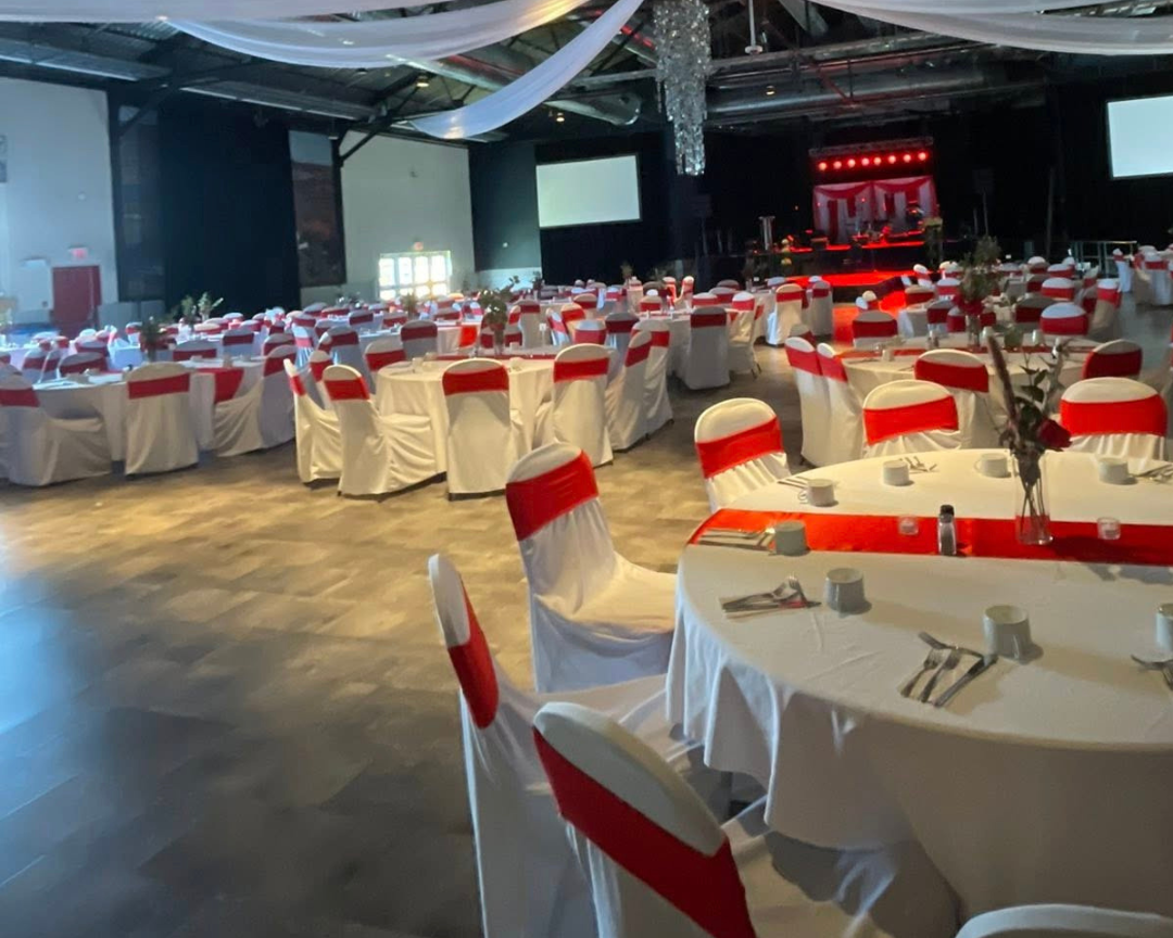 Large indoor event space set for a community gathering, with round tables dressed in white linens, red chair sashes, and a stage prepared for a cultural event.