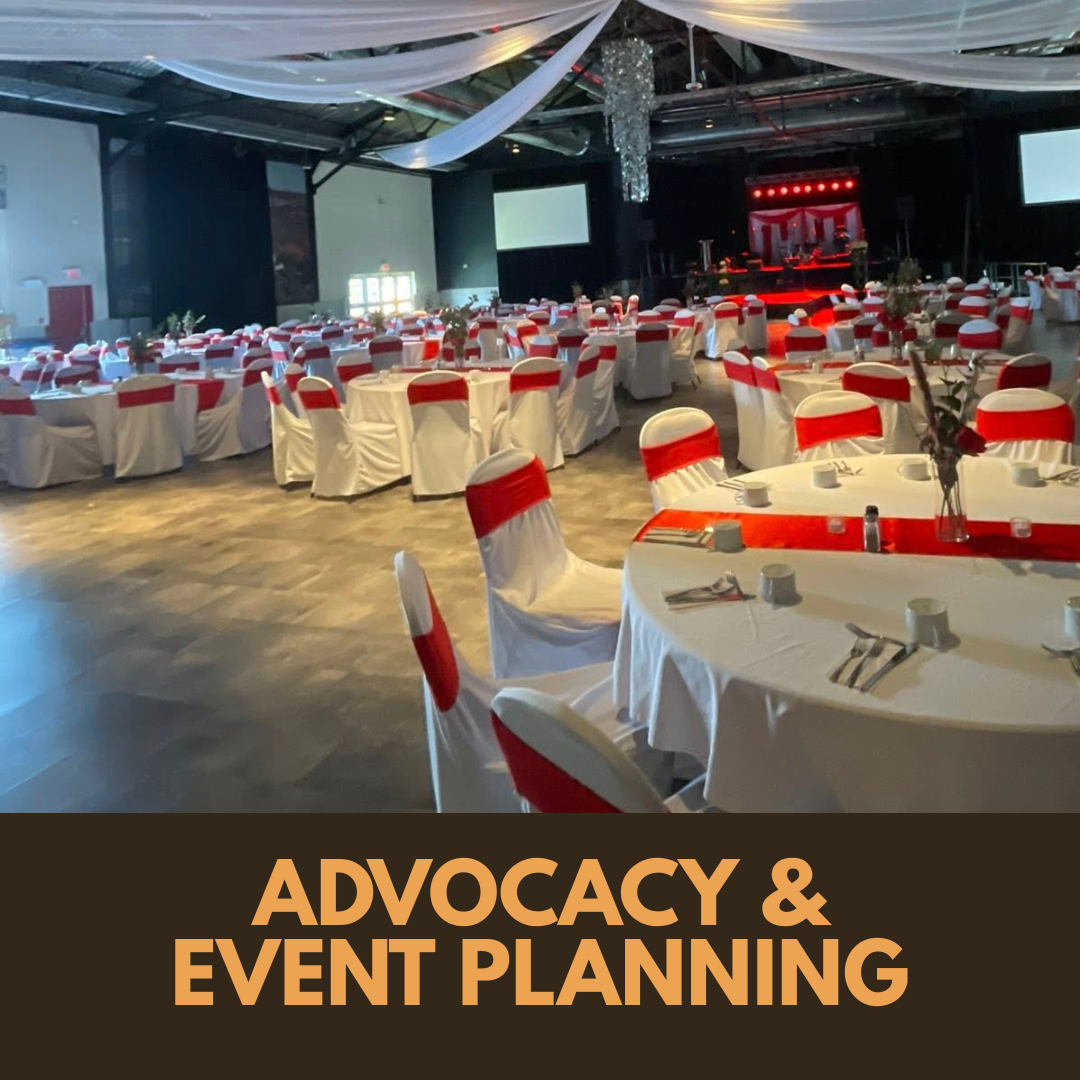 Large indoor event space set for a community gathering, with round tables dressed in white linens, red chair sashes, and a stage prepared for a cultural event.