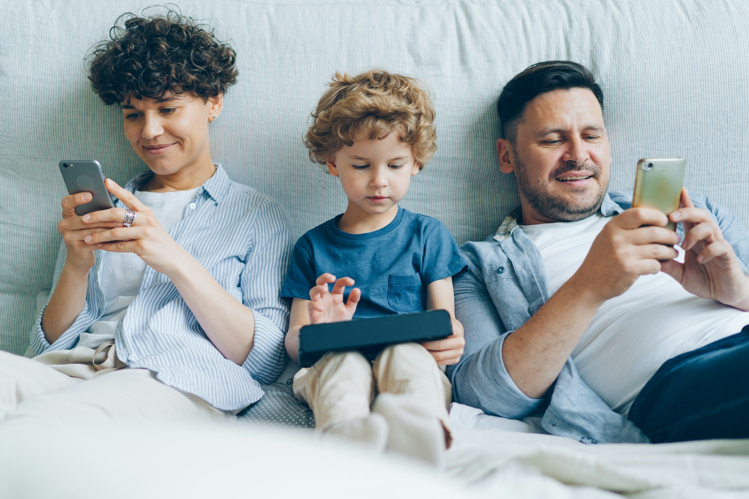 A family of three sitting on a bed, each using a smartphone or tablet. The woman on the left has short, curly hair and is smiling at her phone. The young boy in the middle is focused on his tablet, with a neutral expression. The man on the right has short dark hair and a beard, and is looking at his phone with a slight smile.