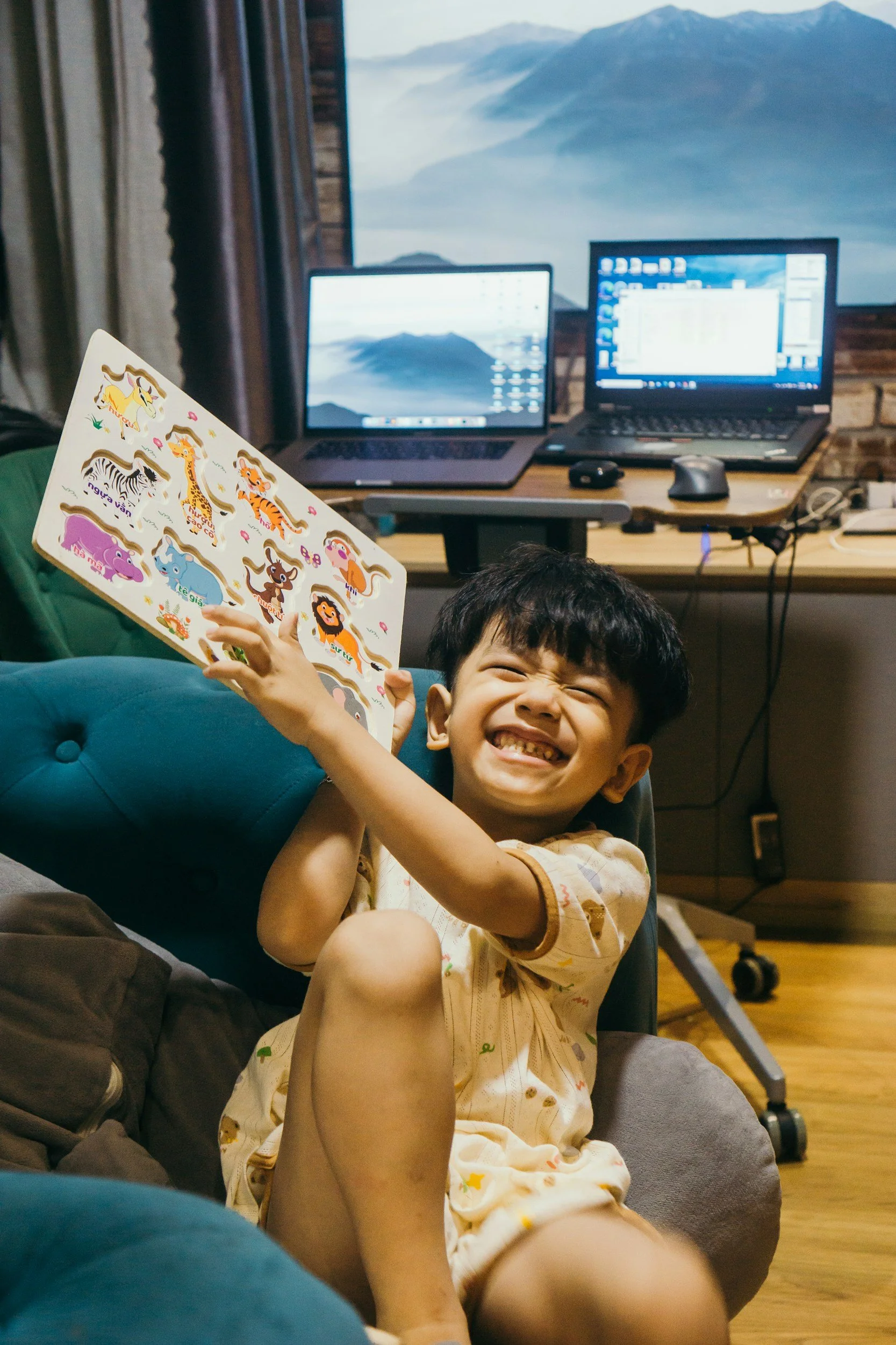 A young boy with a big smile sitting on a couch, holding a sticker book with animal stickers, in a room with two laptops and a mountain landscape on the screen in the background.