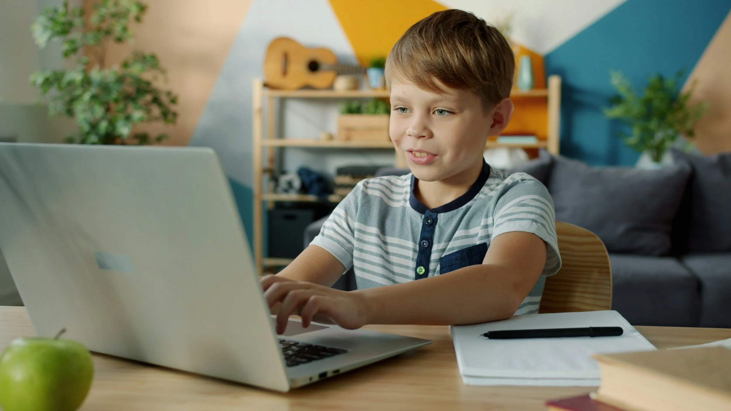 A young boy with short, light brown hair working on a laptop at a desk in a colorful room. There is a green apple, a notebook with a pen, and a book on the desk. In the background, there is a wooden shelf with a guitar, potted plants, and a gray couch.