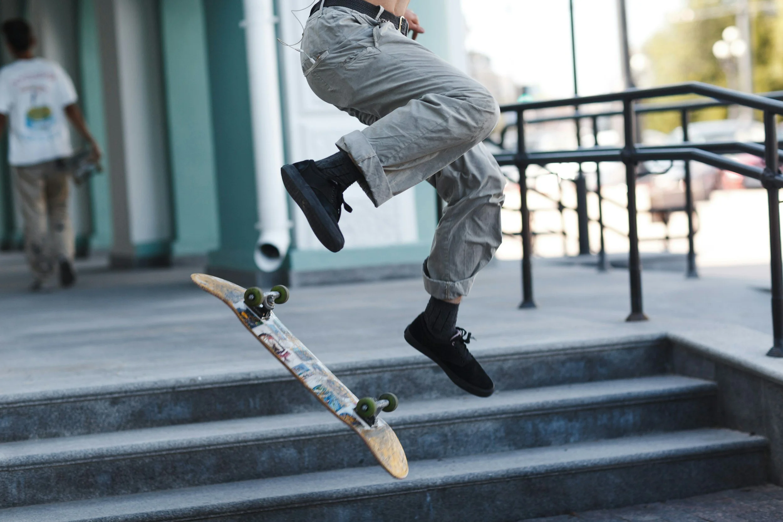 Person in gray pants and black shoes performing a kickflip on a skateboard descending stairs outdoors.