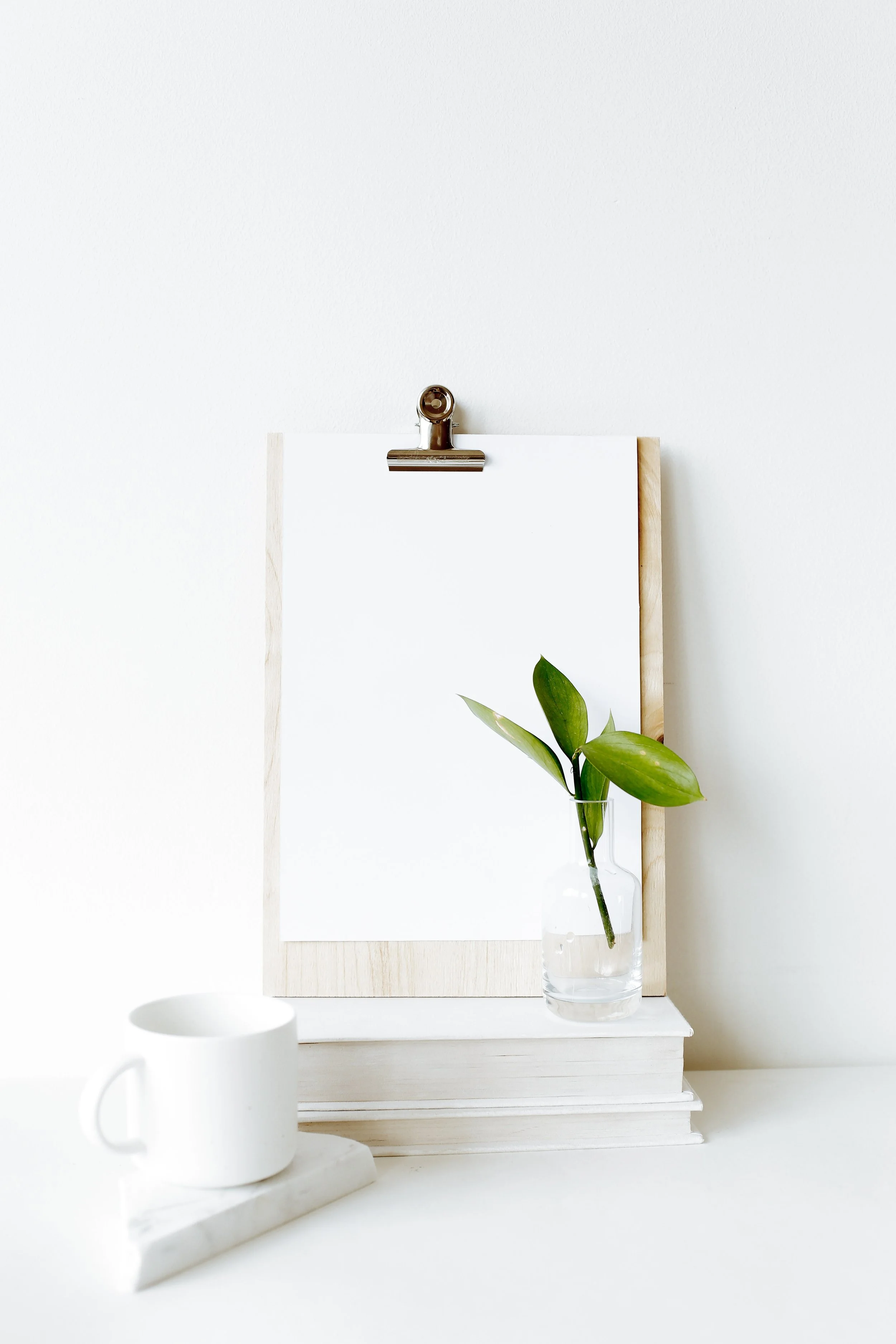 Minimalist scene with a blank white paper on a clip, a small glass vase with green leaves, and a white coffee mug on a marble tray against a white wall.
