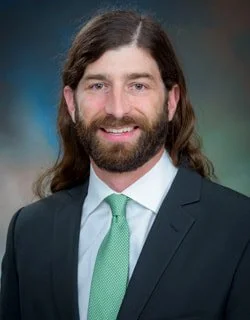 Professional man with long hair and beard, wearing a dark suit, light green tie, and white shirt, smiling against a blurred blue background.