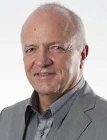A portrait of an older man with short white hair, wearing a light gray blazer and a darker gray shirt, smiling at the camera.