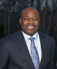 A smiling African American man dressed in a dark suit, white shirt, and patterned tie outdoors.