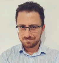 A man with glasses, a beard, and curly dark hair, wearing a light blue striped shirt, looking slightly downward