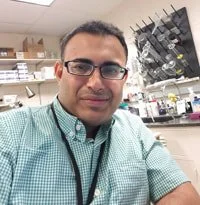 A man with glasses wearing a light green checkered shirt taking a selfie in a laboratory or pharmacy setting with shelves of supplies and equipment in the background.