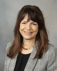 Smiling woman with shoulder-length brown hair, wearing a gray checkered blazer and a black top, against a neutral background.