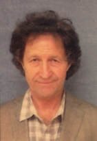 Portrait of a man with curly dark hair, wearing a light-colored, checkered collared shirt, facing forward against a plain background.