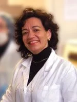 A woman with curly dark hair smiling, wearing a white lab coat and black turtleneck, in a professional setting.