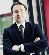 A man in a suit with arms crossed, standing indoors near a window.